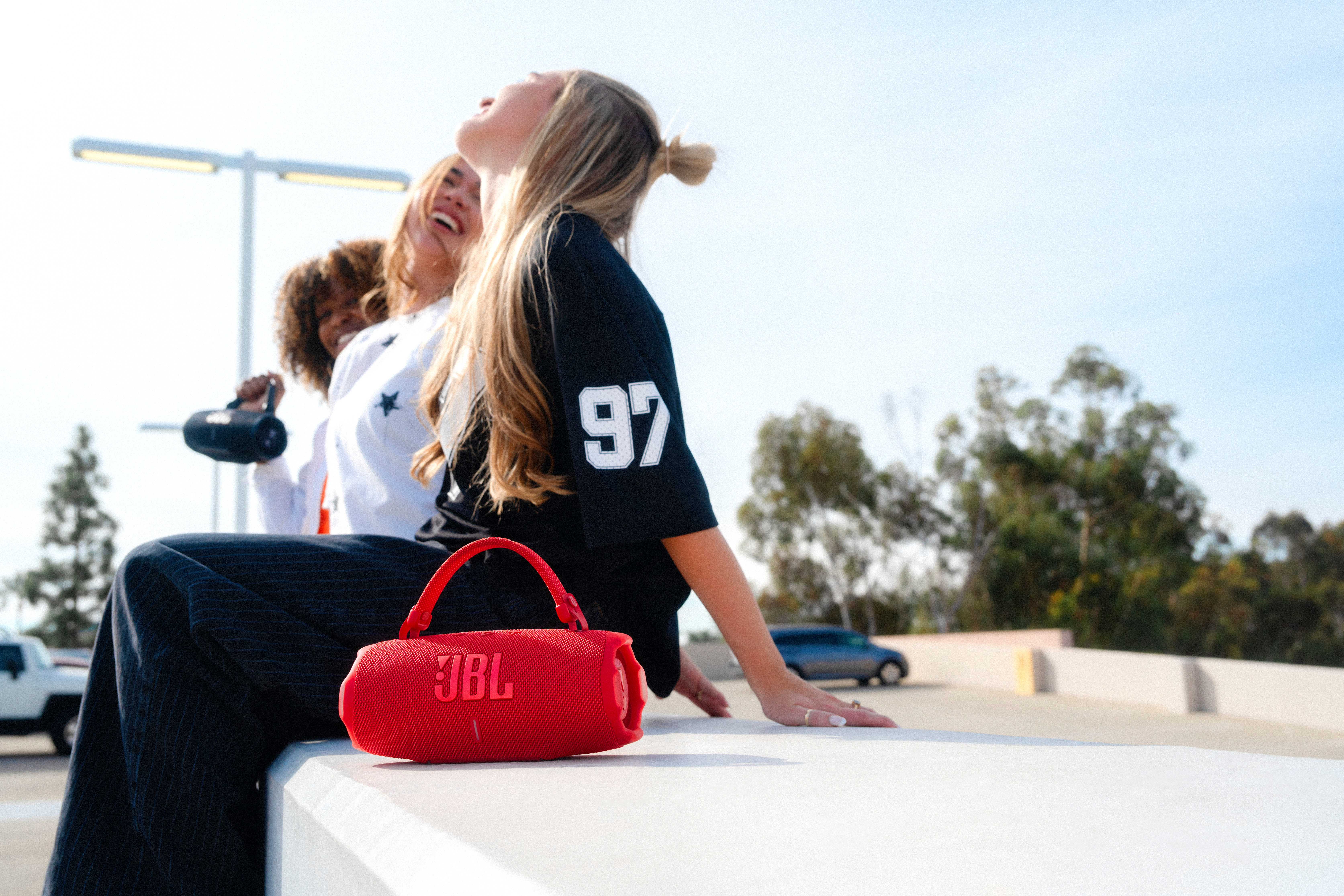 Trois femmes assises, une tenant une enceinte JBL rouge. Fond de paysage urbain.