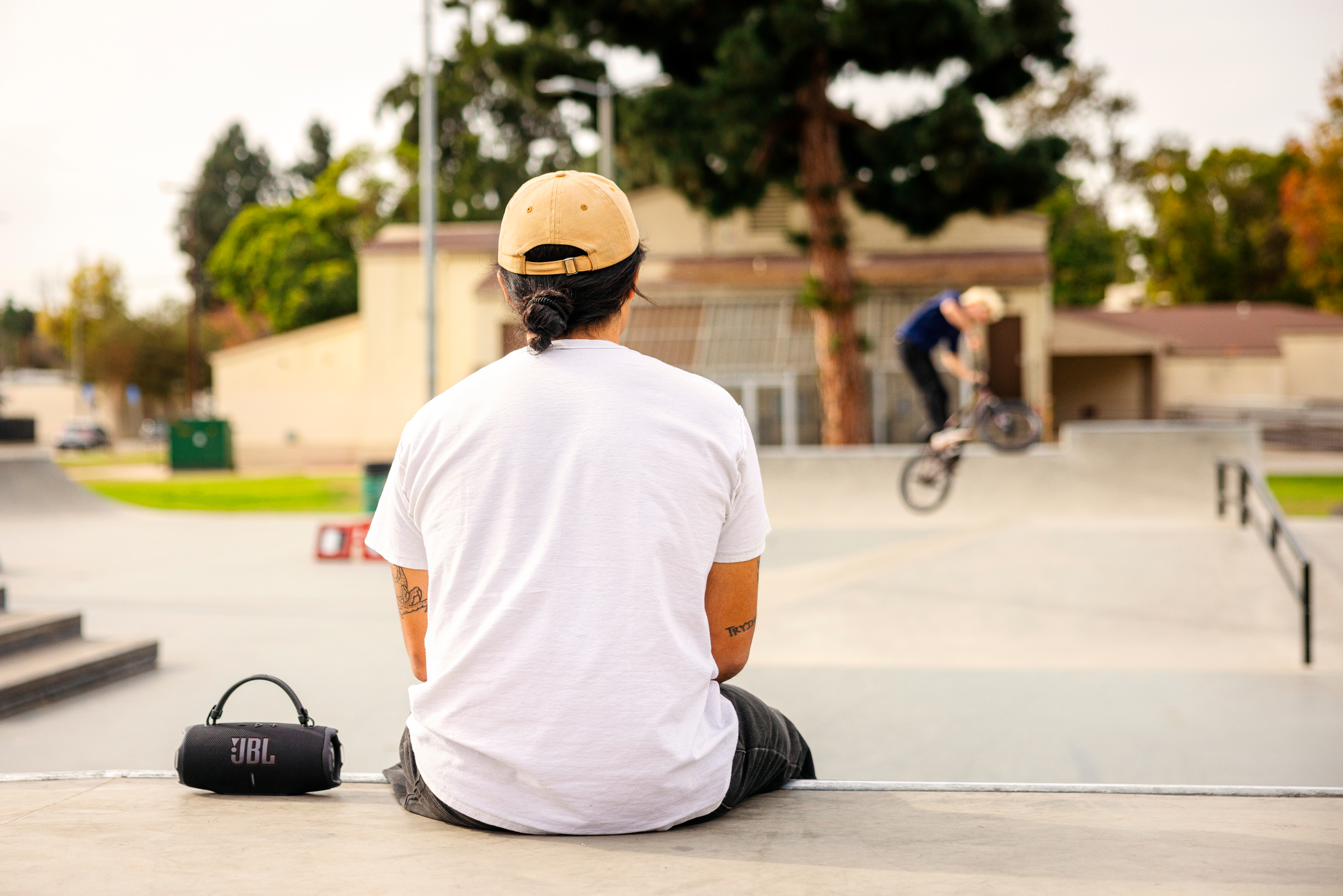 Mann sitzt, schaut BMX-Action. JBL-Lautsprecher am Boden. Skatepark-Szene.