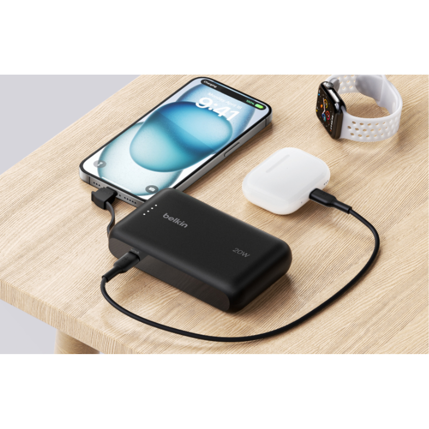 Une batterie externe Belkin charge un téléphone et des AirPods sur une table en bois. Le téléphone se charge.