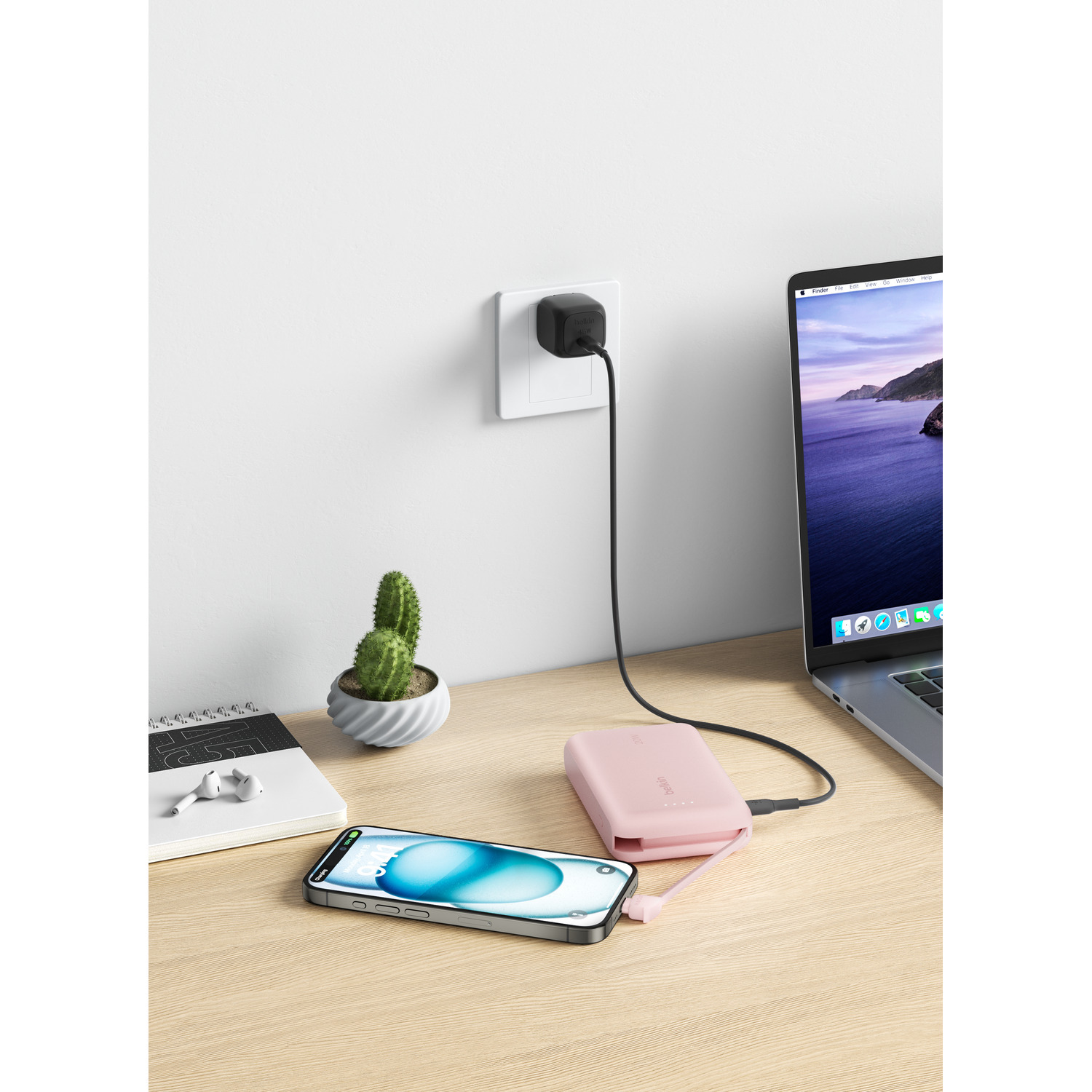Batterie externe rose chargeant téléphone sur bureau. Ordinateur portable, cactus et chargeur visibles. Mur blanc.