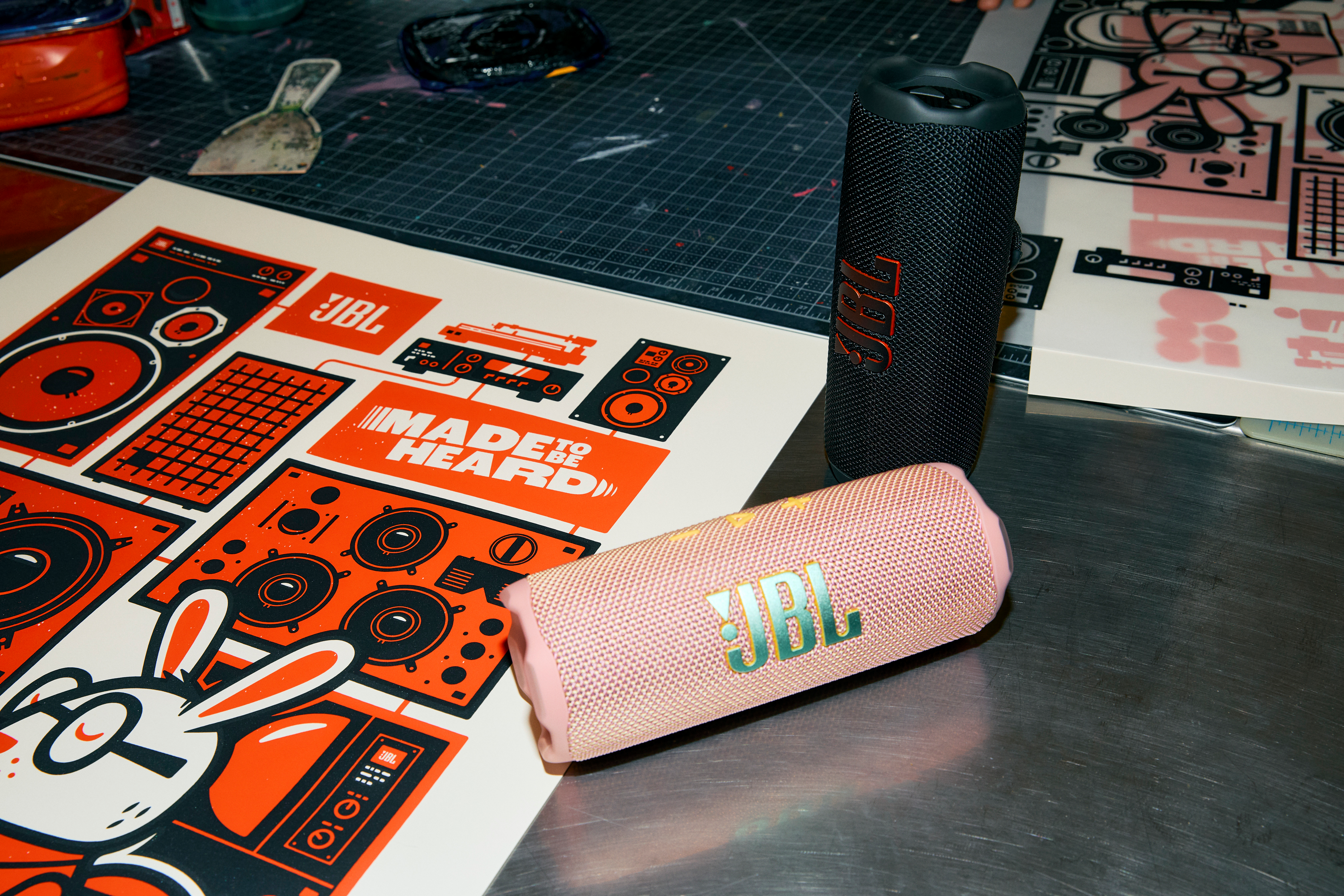 Zwei JBL Flip 7 Lautsprecher und Grafik. Ein Lautsprecher ist schwarz, einer rosa. Auf einem Tisch.