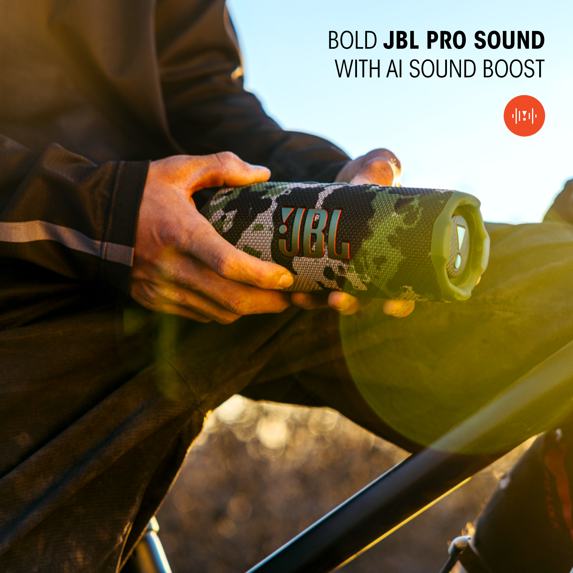 Person hält JBL Flip 7 Lautsprecher. Camouflage-Muster. Fahrrad im Hintergrund, sonniger Tag.