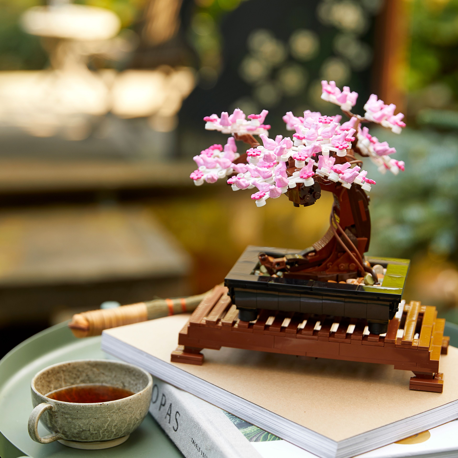 Lego bonsai boom op tafel. Een lego bonsai boom op een houten standaard met een kopje en een boek.