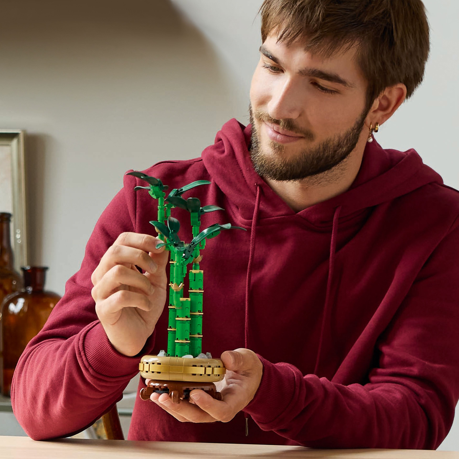 A man with a beard is holding a Lego Lucky Bamboo plant. He is smiling. The background is a room.