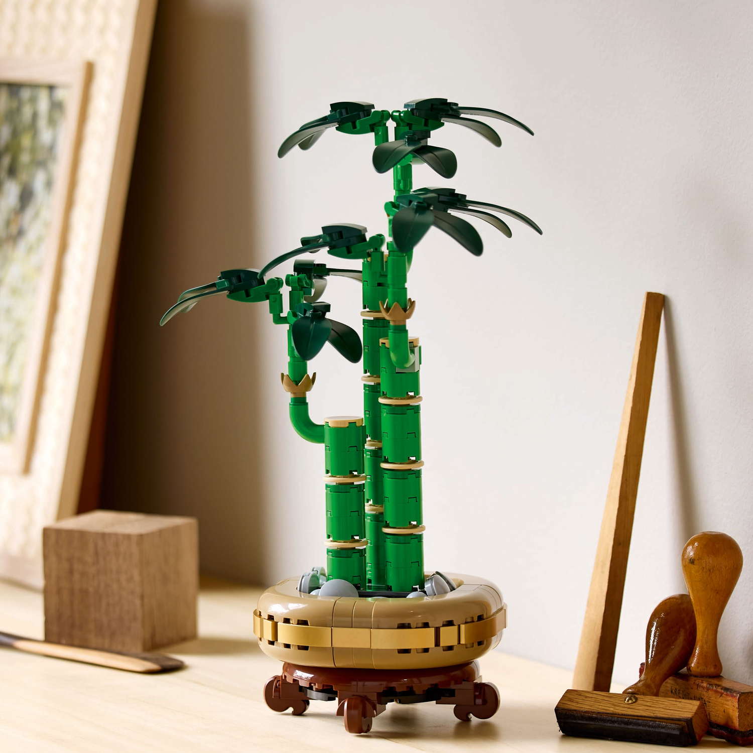 Lego Lucky Bamboo plant in a pot on a wooden table. The background includes a framed picture and wooden elements.