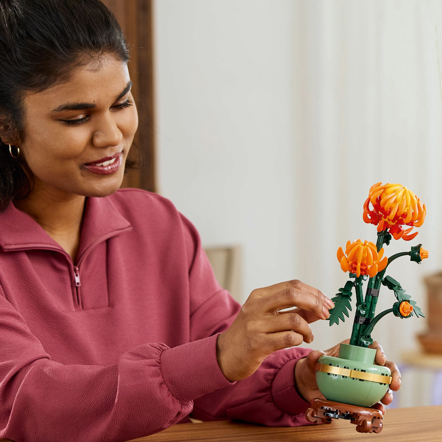 A woman is holding a Lego Chrysanthemum plant, looking at it with a smile. The background is a room.