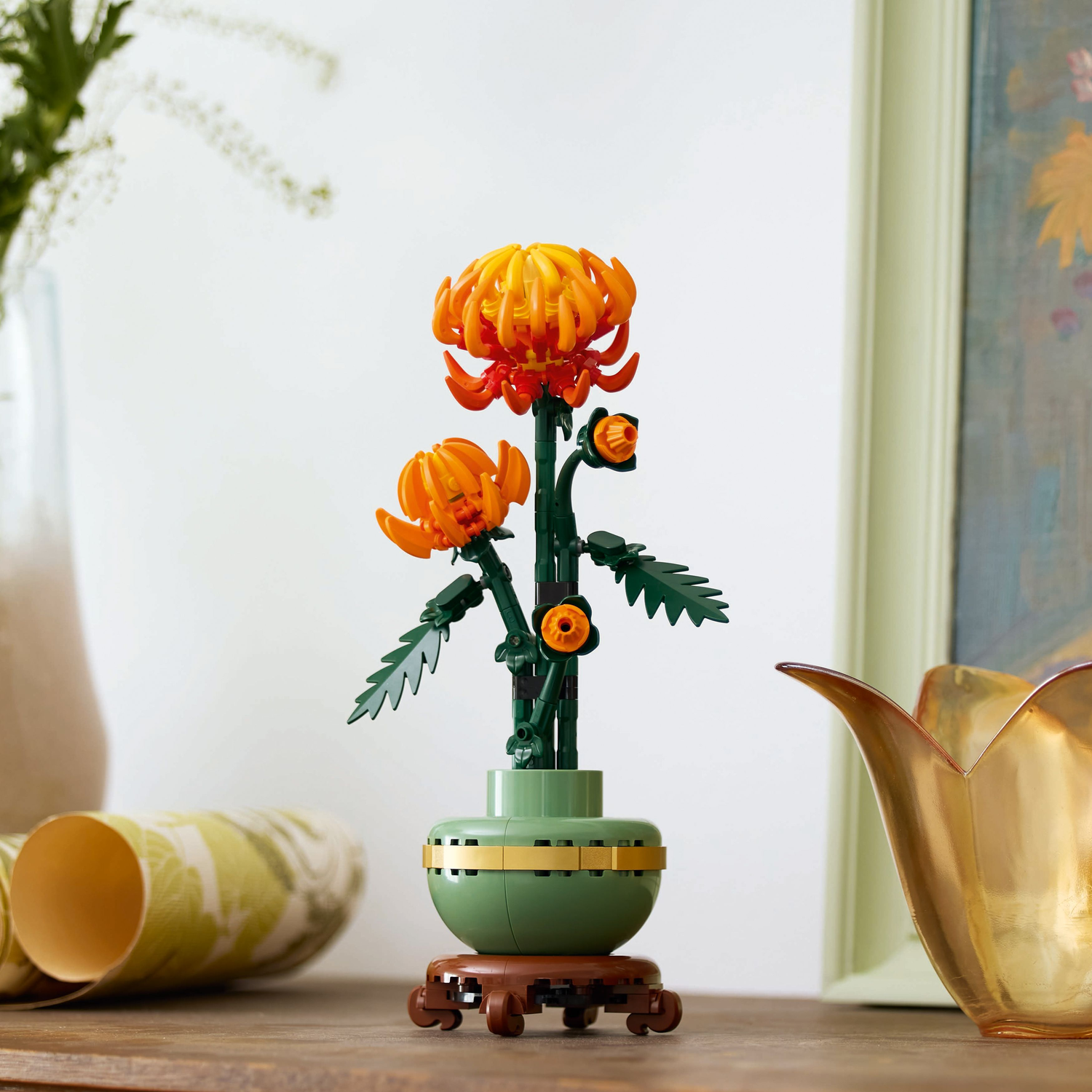 Lego Chrysanthemum in a pot on a wooden table. Background includes a golden vase and a framed painting.