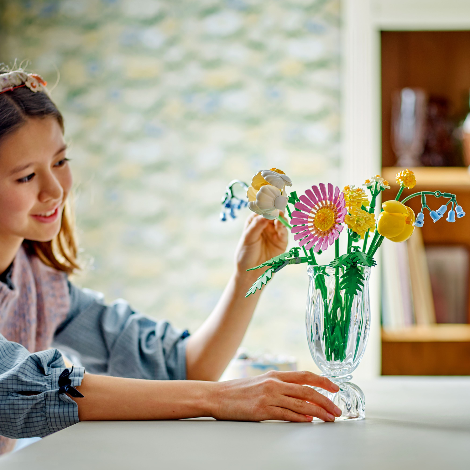 A girl is arranging a Lego flower bouquet in a glass vase. The bouquet features pink, yellow, and blue Lego flowers.