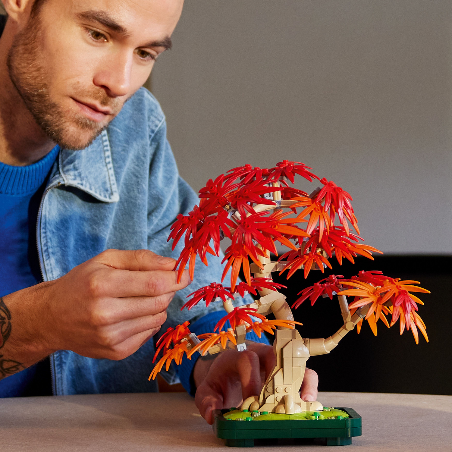 A man is holding a Lego bonsai tree model with red and orange leaves. He is focused on the Lego model.