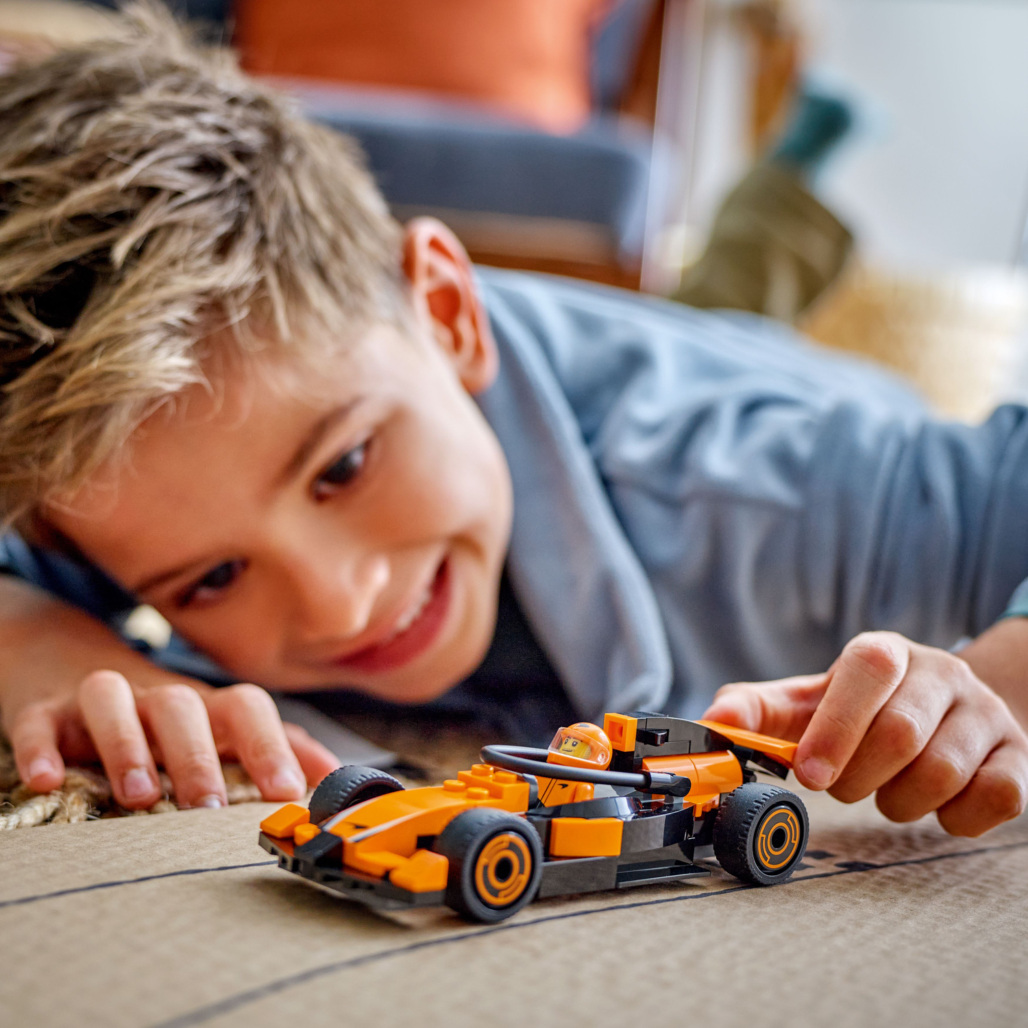 A boy smiles at a Lego race car, lying on a patterned surface.