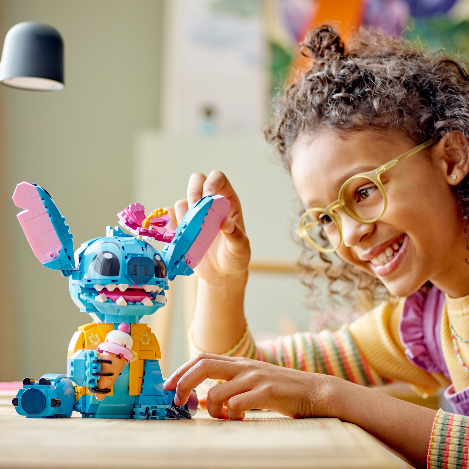 Meisje speelt met Lego Stitch. Stitch houdt ijs vast. Geel shirt, bril. Achtergrond: kunst.