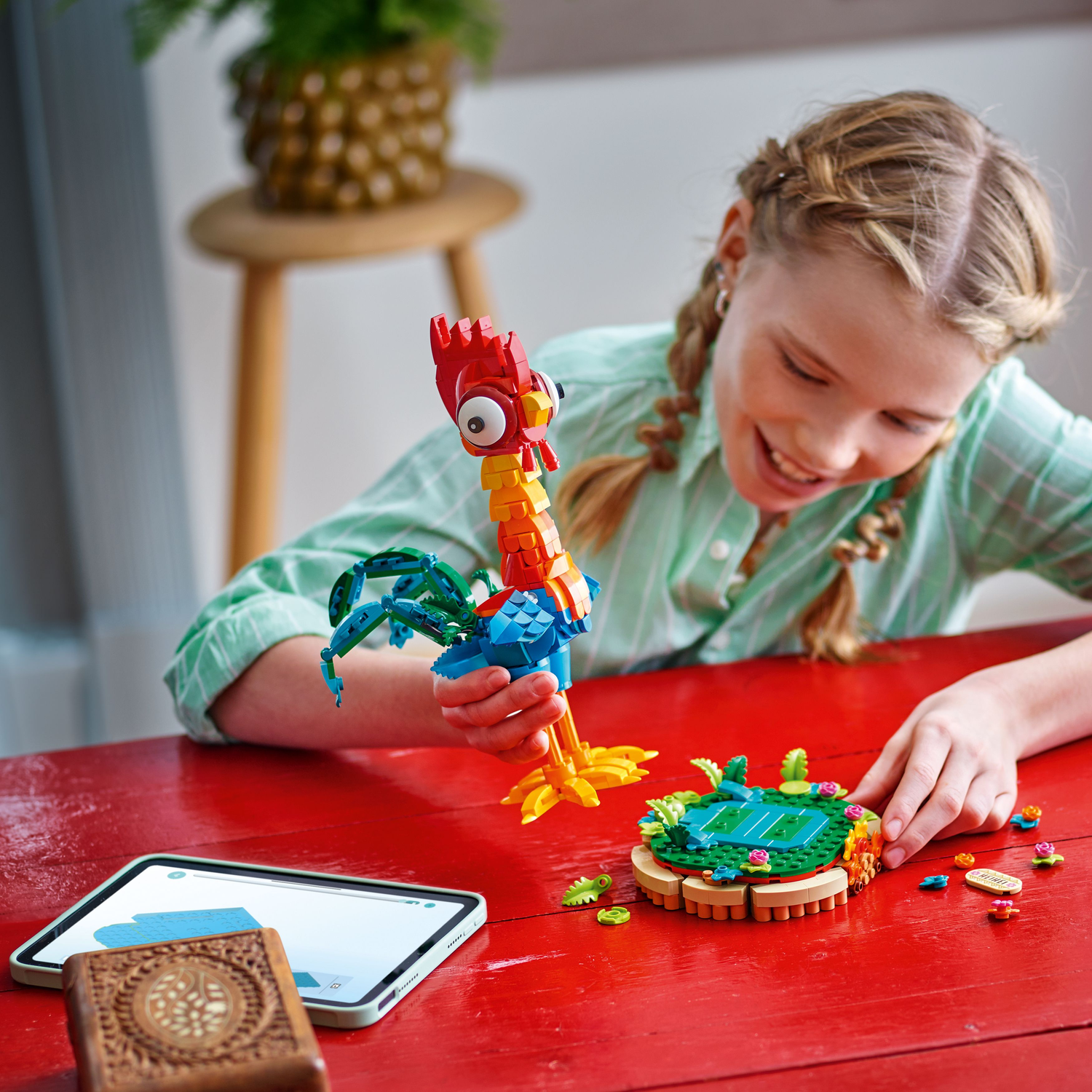 Een meisje speelt met een LEGO-haanmodel, naast een tablet en een kleine basis met bloemen.
