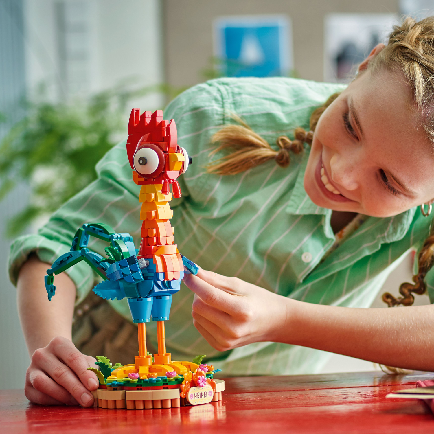 Een meisje interageert met een LEGO-haanmodel op een rode tafel. Het meisje glimlacht.
