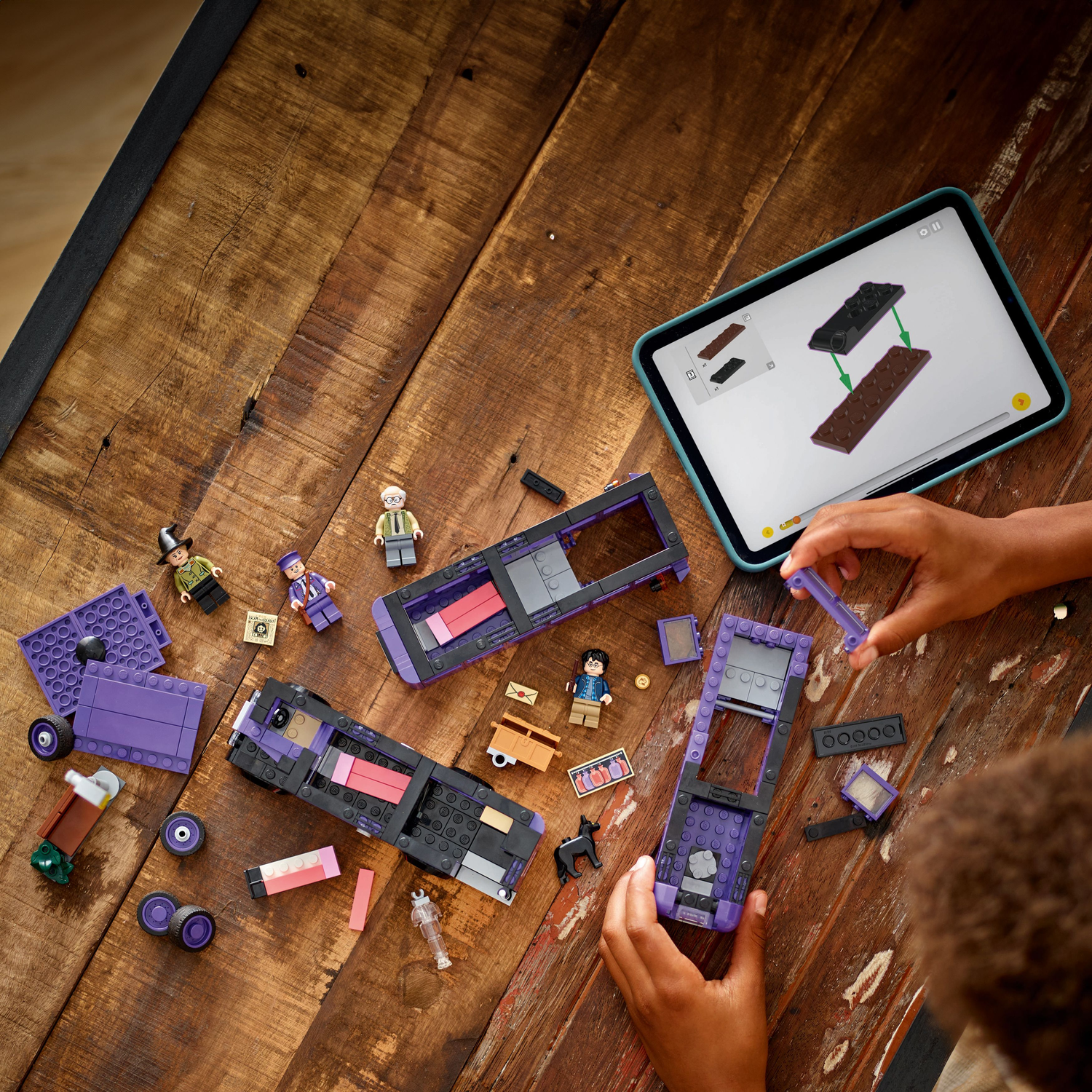 Hands of a child building a LEGO Knight Bus model, with minifigures and a tablet showing instructions.