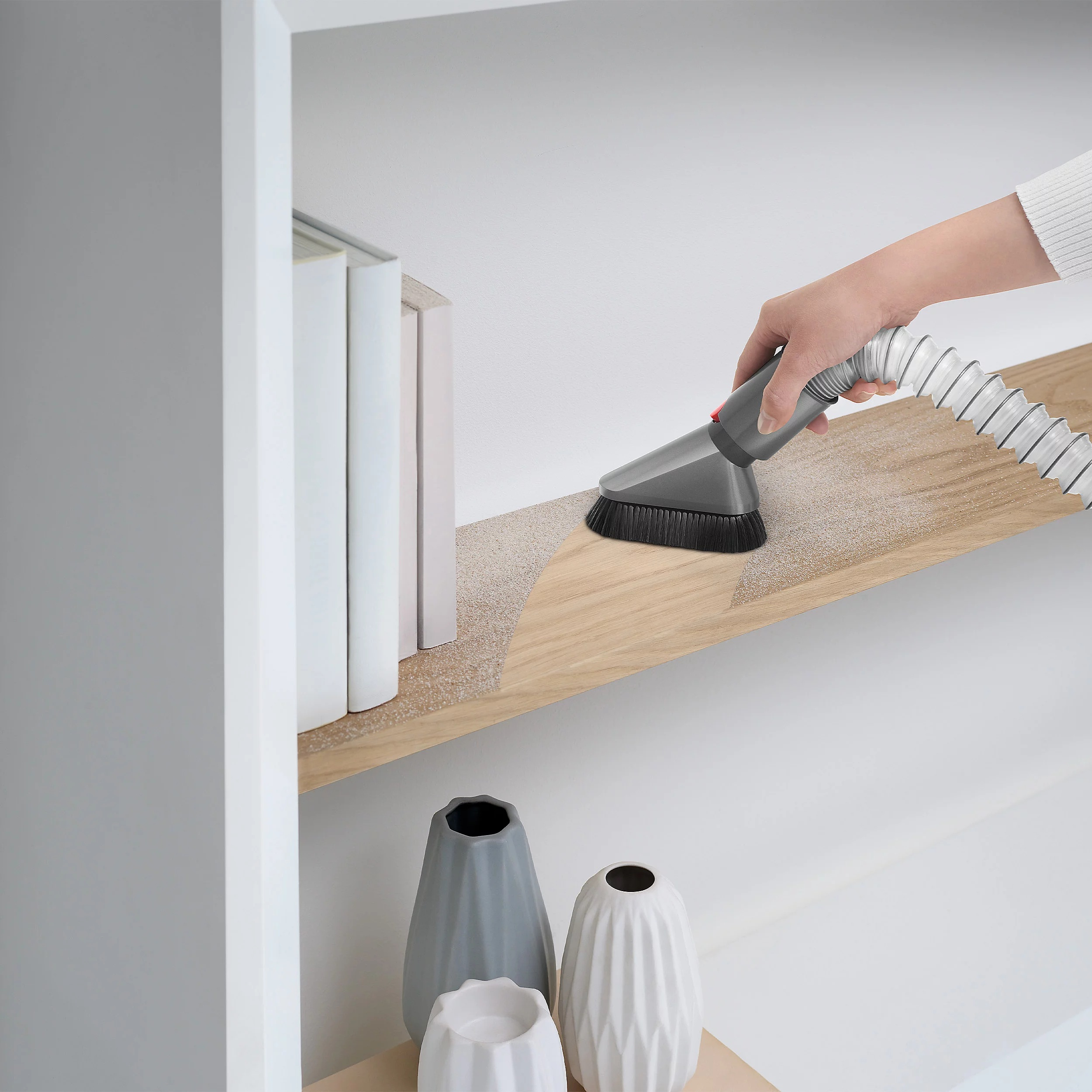 Osoba używająca nasadki do odkurzacza do czyszczenia półki z książkami i wazonami.