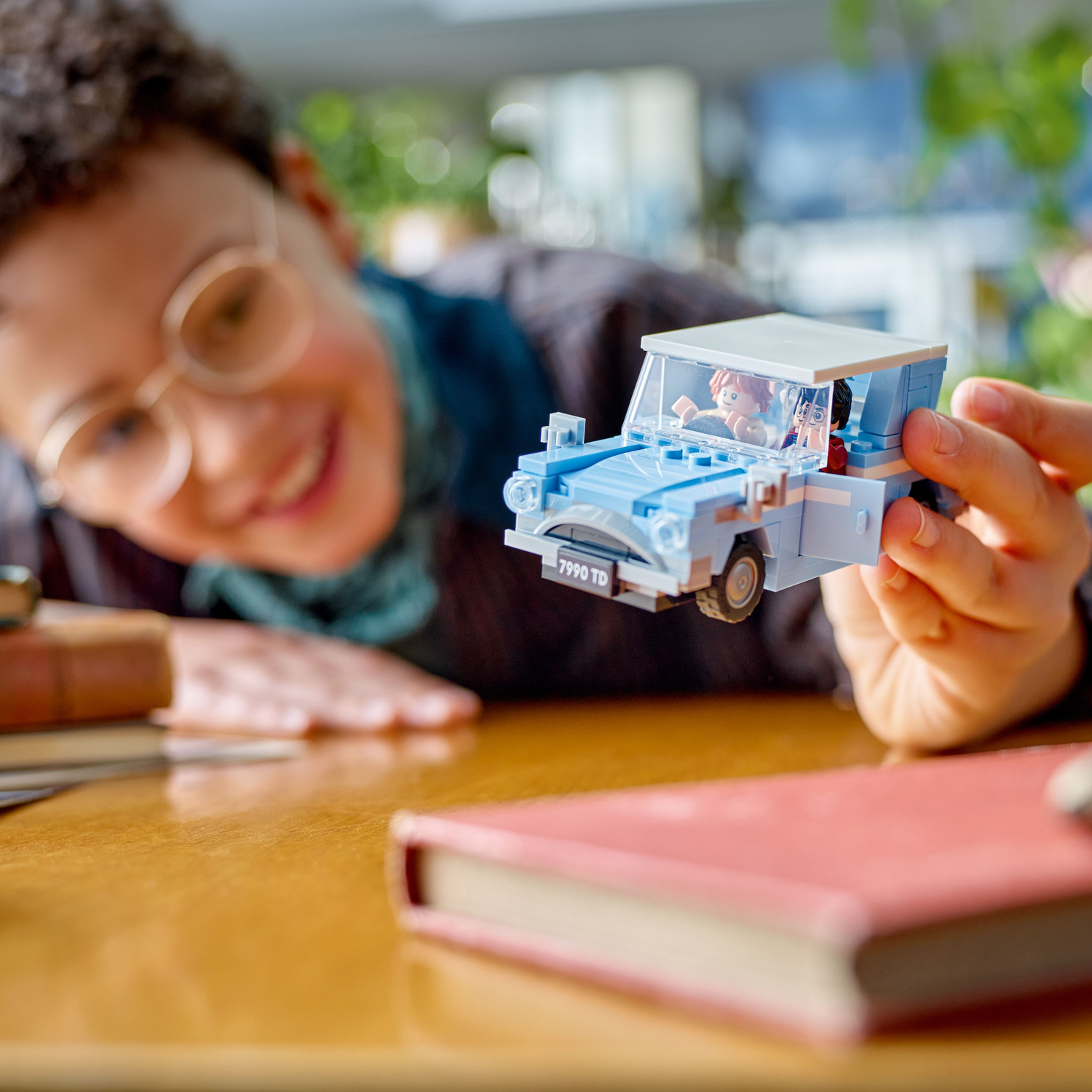 Person smiles, holding a blue Lego car. A red book is on a wooden table.