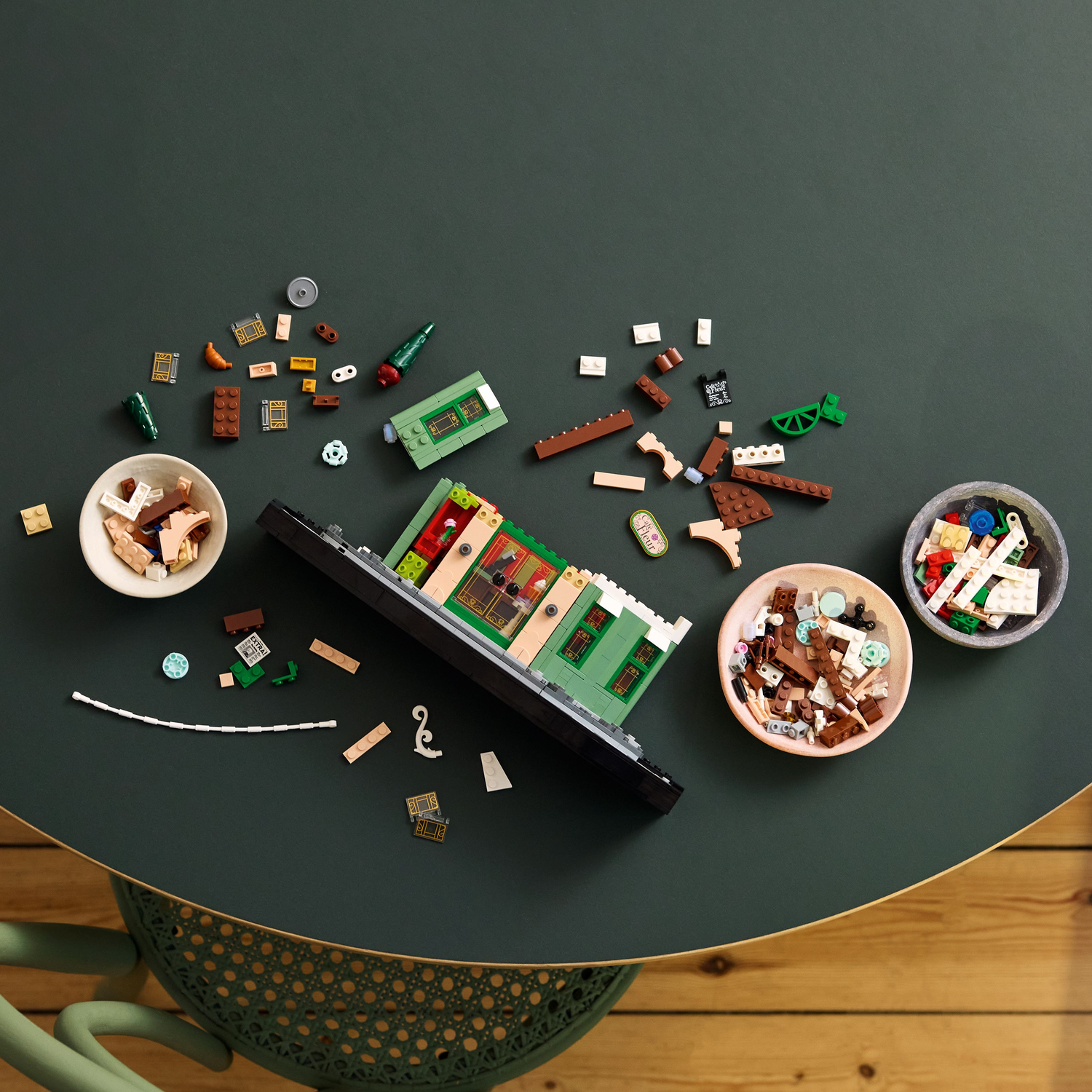 LEGO bricks are scattered on a green table with a partially built cafe model and bowls.