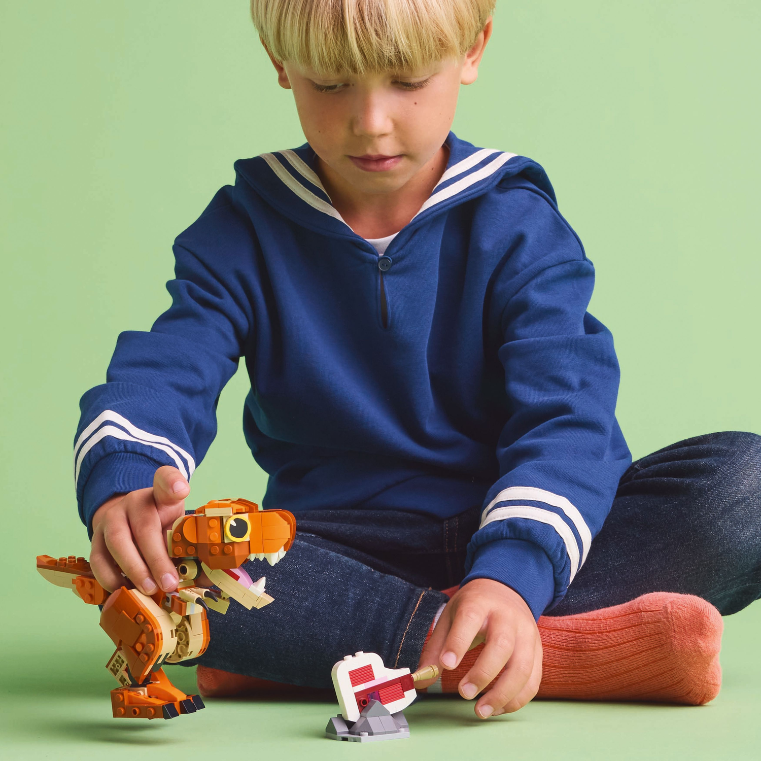 Een jongen speelt met een oranje Lego T-rex dinosaurus speelgoed en een wit taart speelgoed op groen.