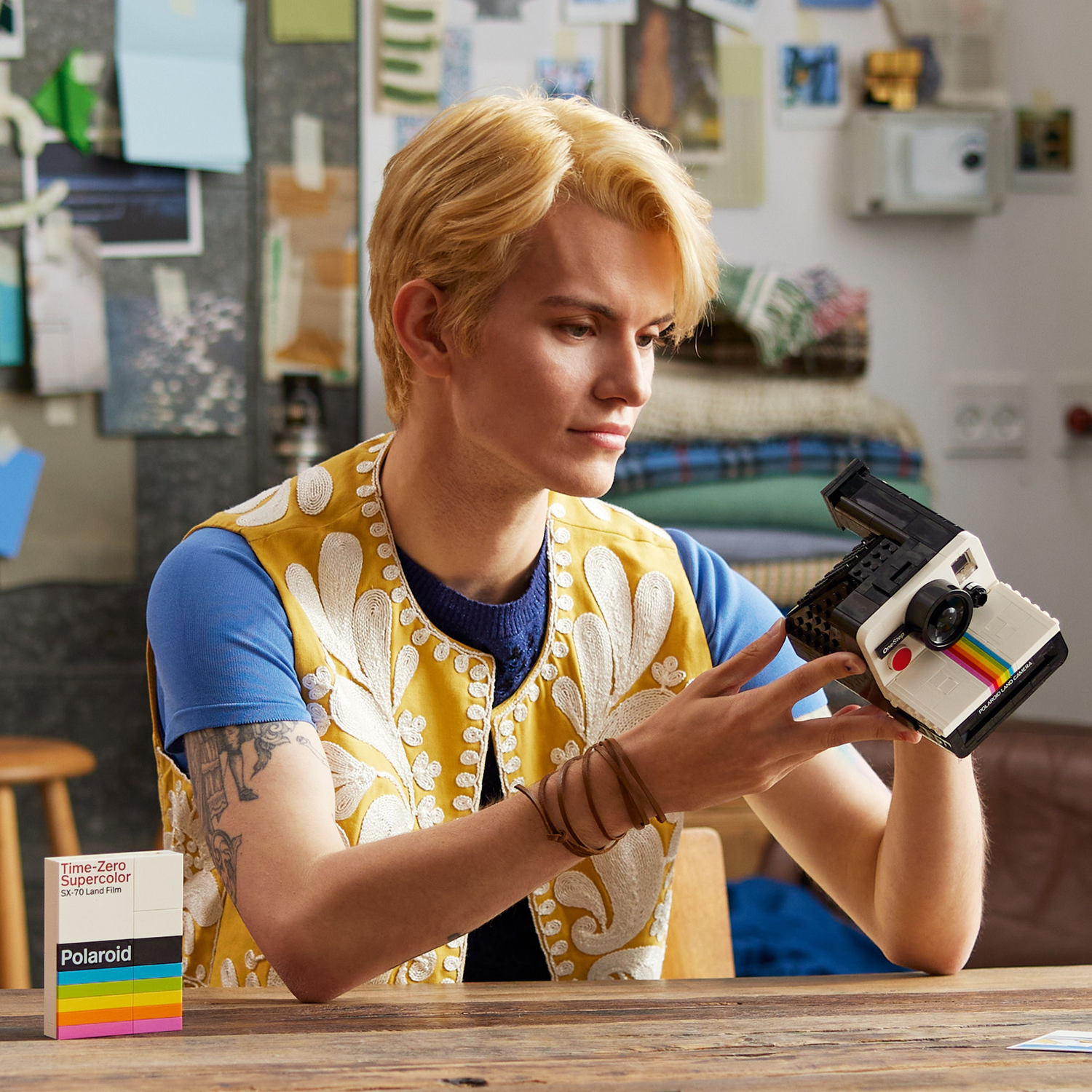 Man met het LEGO Polaroid cameramodel. Blond haar, gele vest, blauw shirt. Foto's en film op tafel.