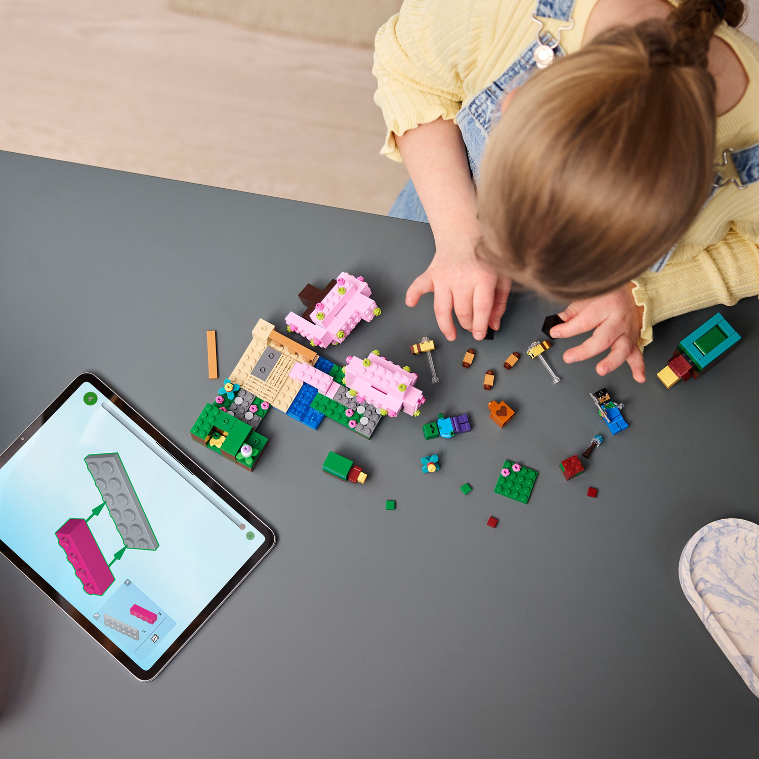 Een kind bouwt een Lego Minecraft set met een tablet met instructies. Set onderdelen en figuren liggen op een tafel.