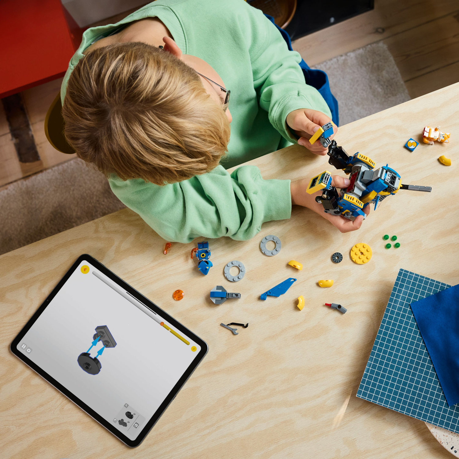 A boy builds a Lego Sonic set. Parts are on the table. A tablet shows instructions.