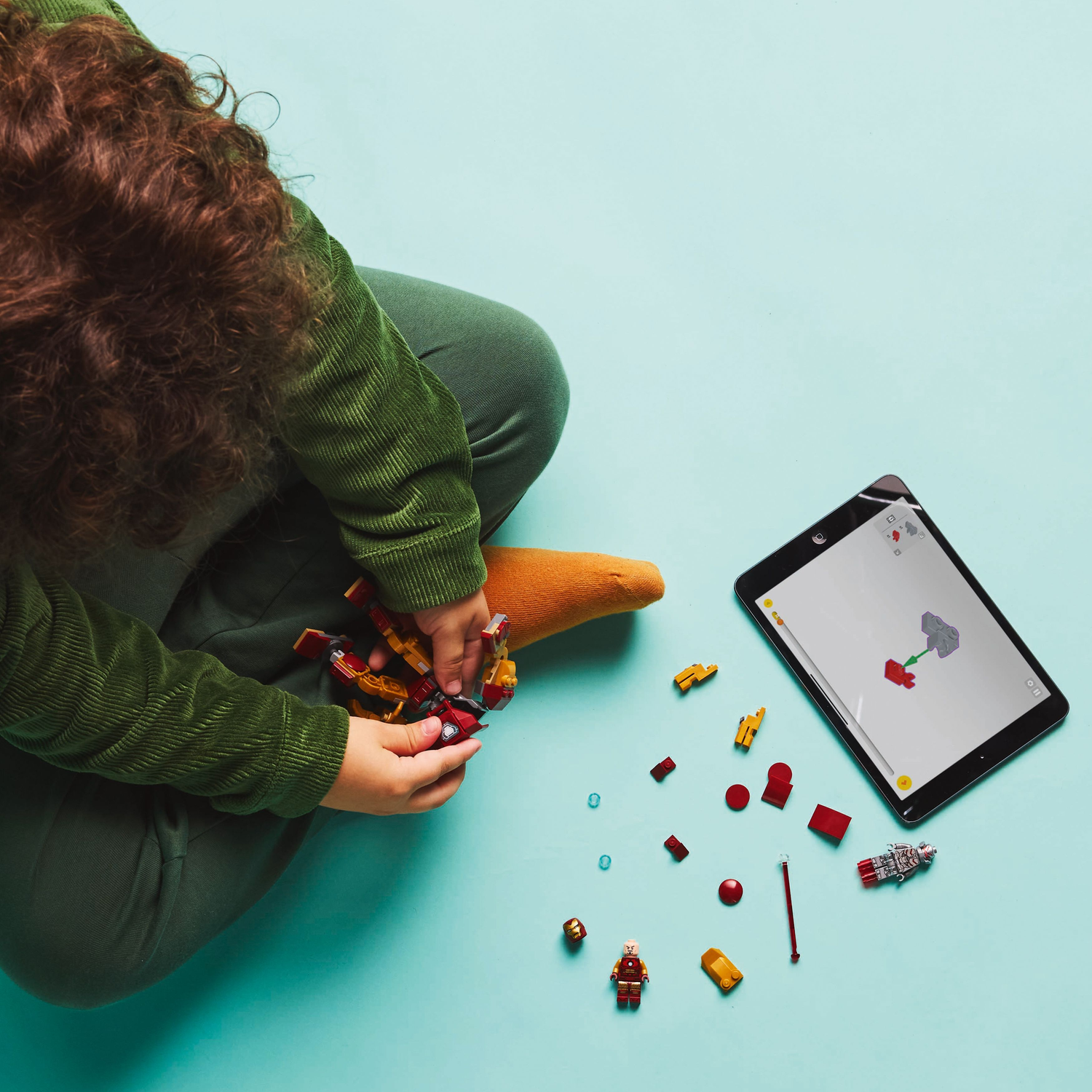 A child sits assembling a Lego Iron Man mech. A tablet displays instructions. Scattered Lego pieces surround.