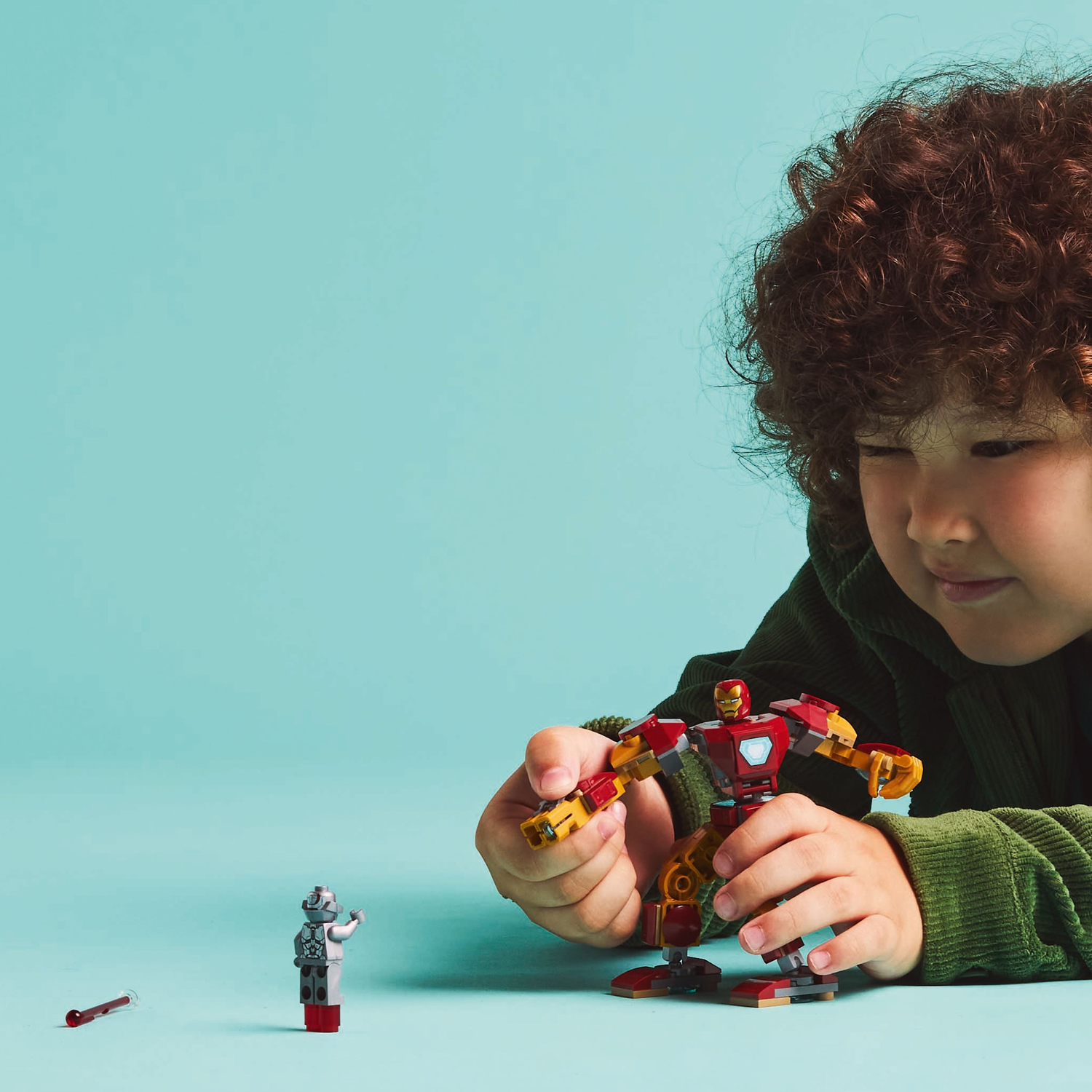 A child holds an Iron Man mech Lego, facing an Ultron Lego figure. A red Lego rod lies on the surface.