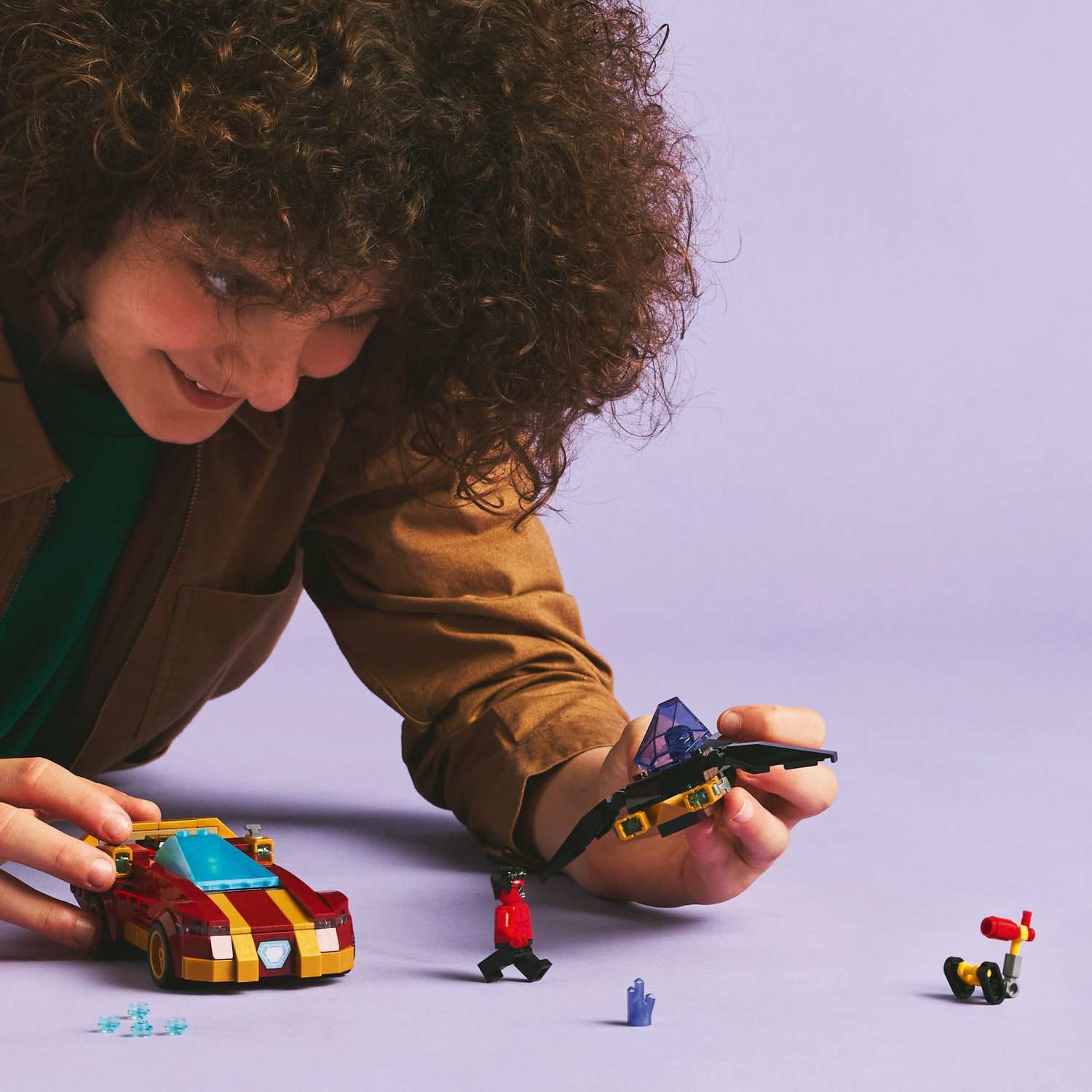 A child is playing with a Lego set, including a car, jet, and figures of Iron Man, Red Hulk, and others.