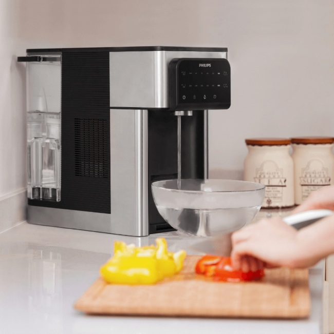 Scena kuchenna: Dystrybutor wody Philips dozuje wodę do miski. Warzywa na blacie.