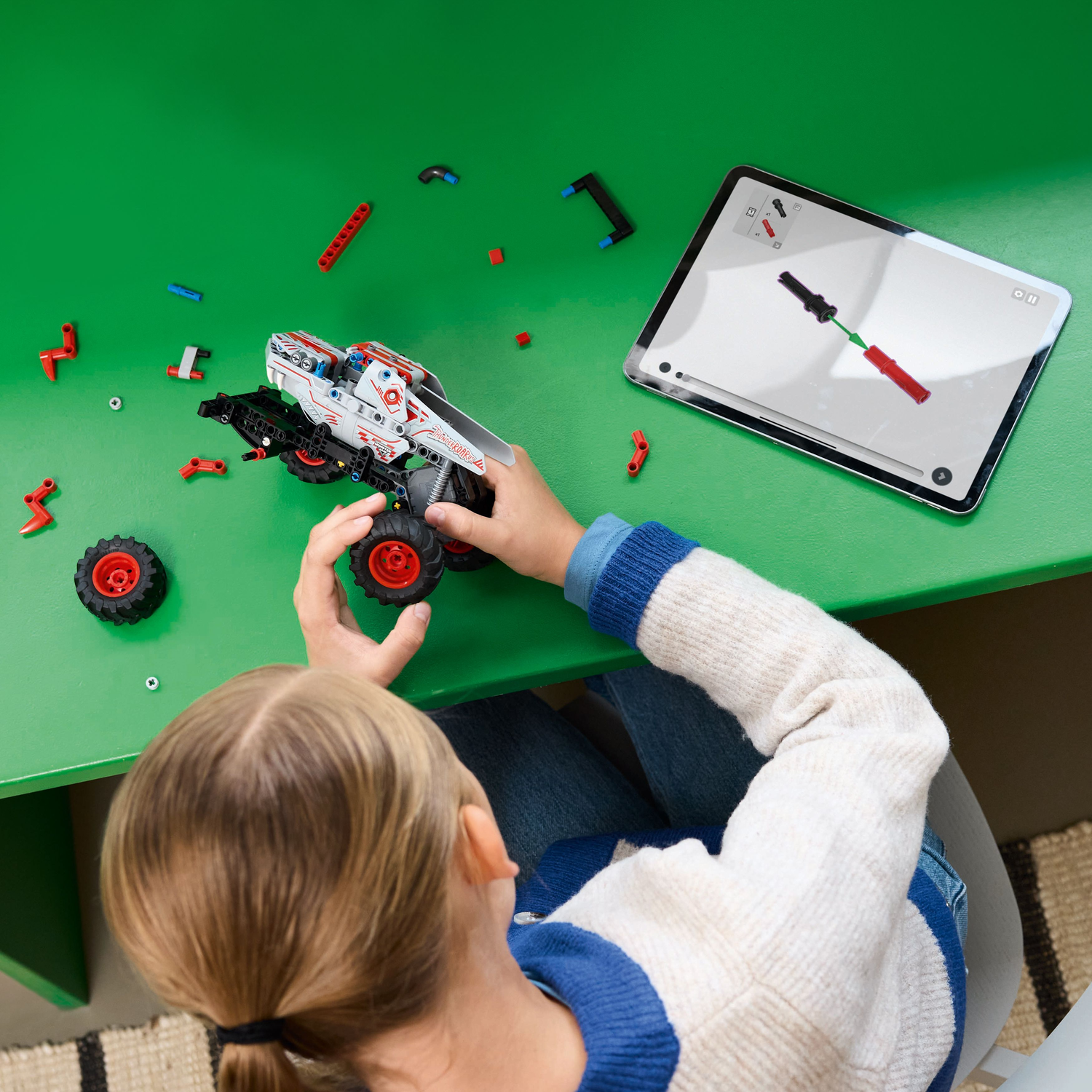 Girl building a gray and red toy monster truck on a green table. Tablet nearby.