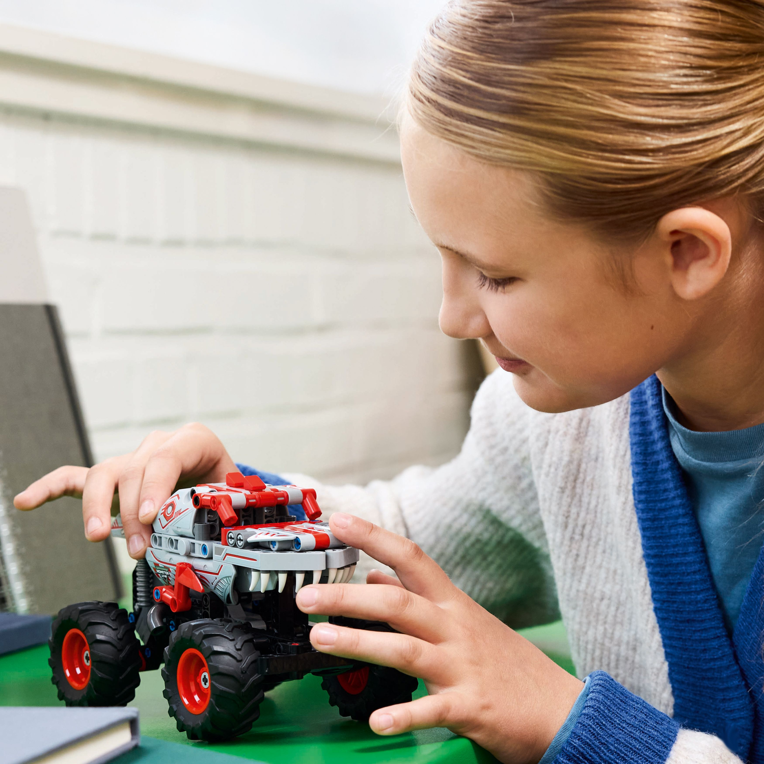 Girl looking at a gray and red toy monster truck on a green table.