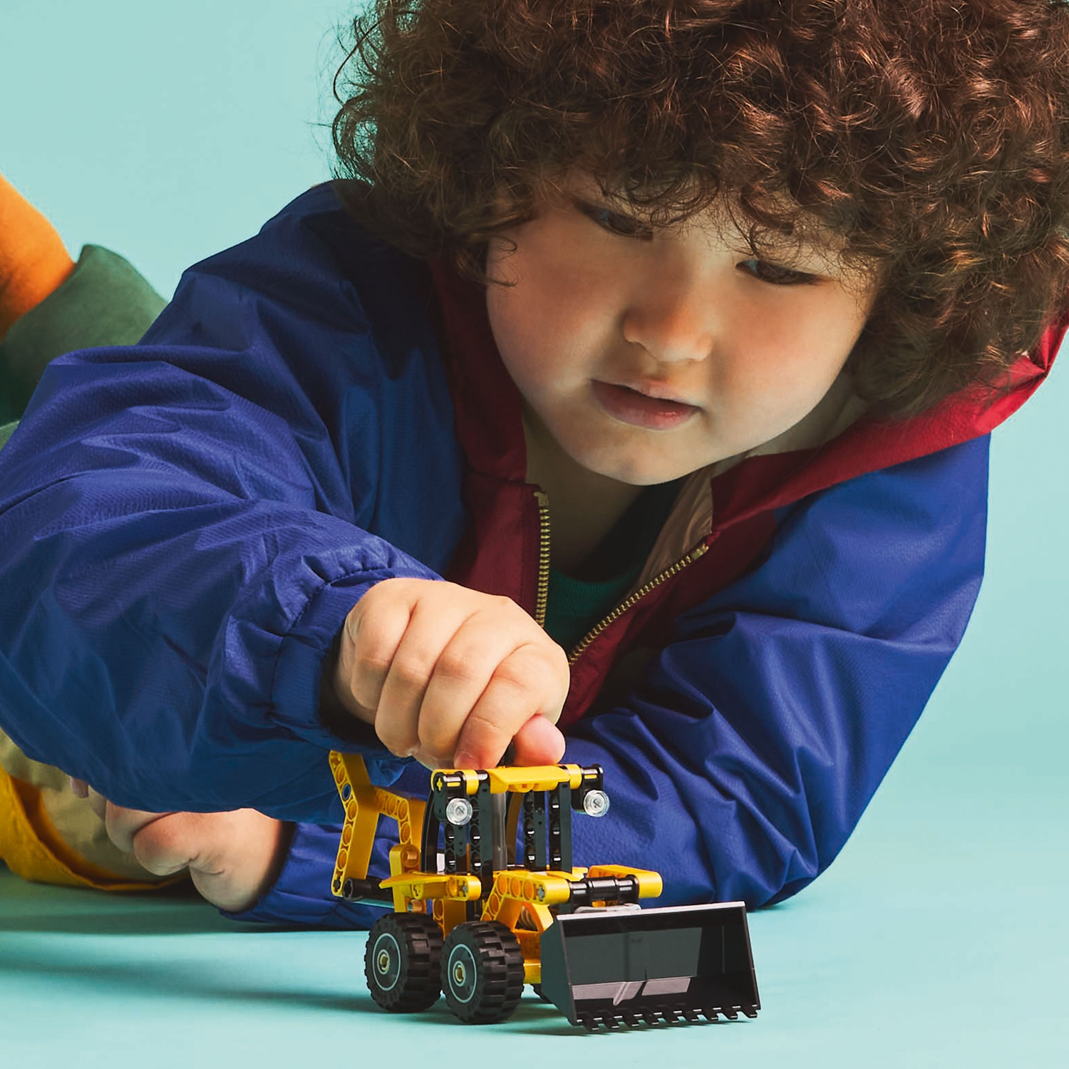 Child holding a small yellow Lego backhoe loader, on a blue background.