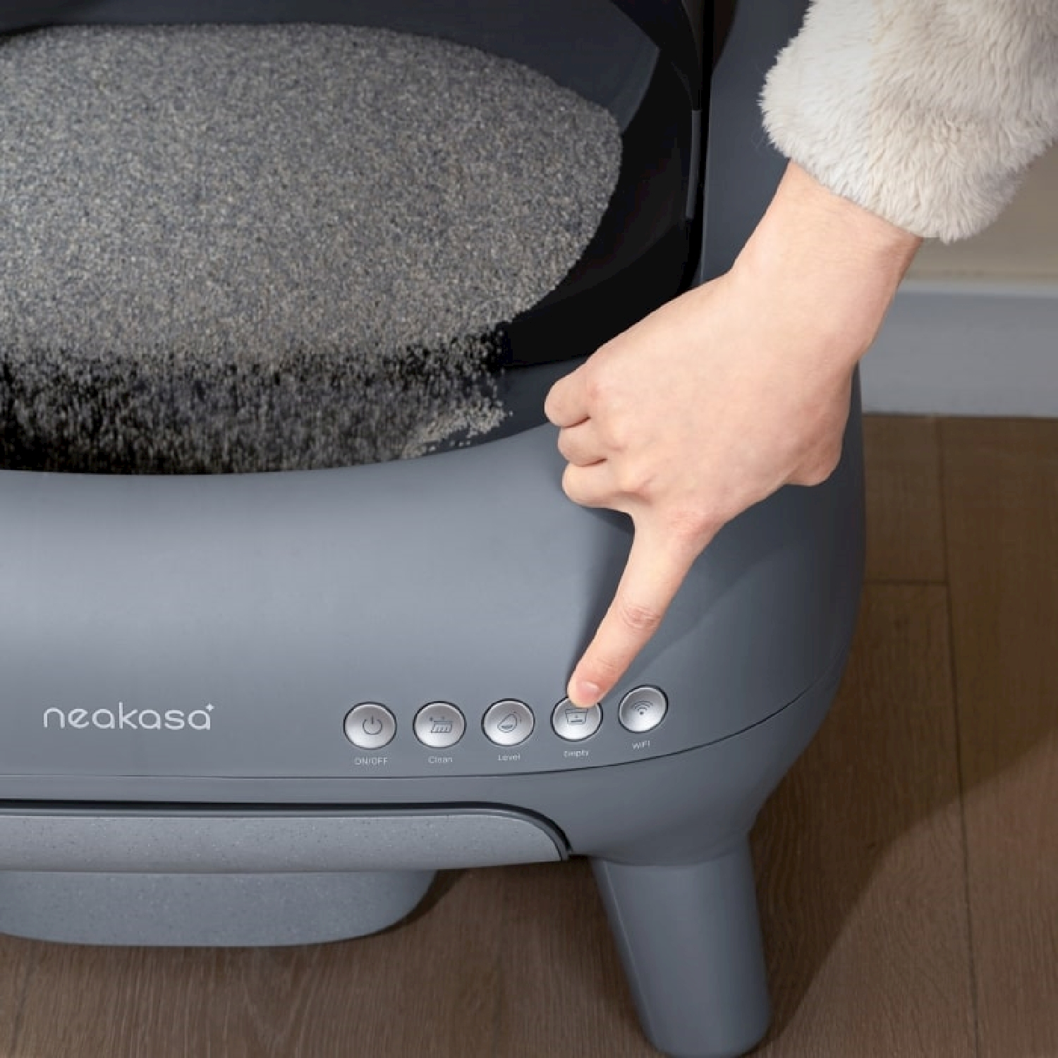 Una mano preme il pulsante 'Sweep' su una lettiera automatica grigia e moderna per gatti.