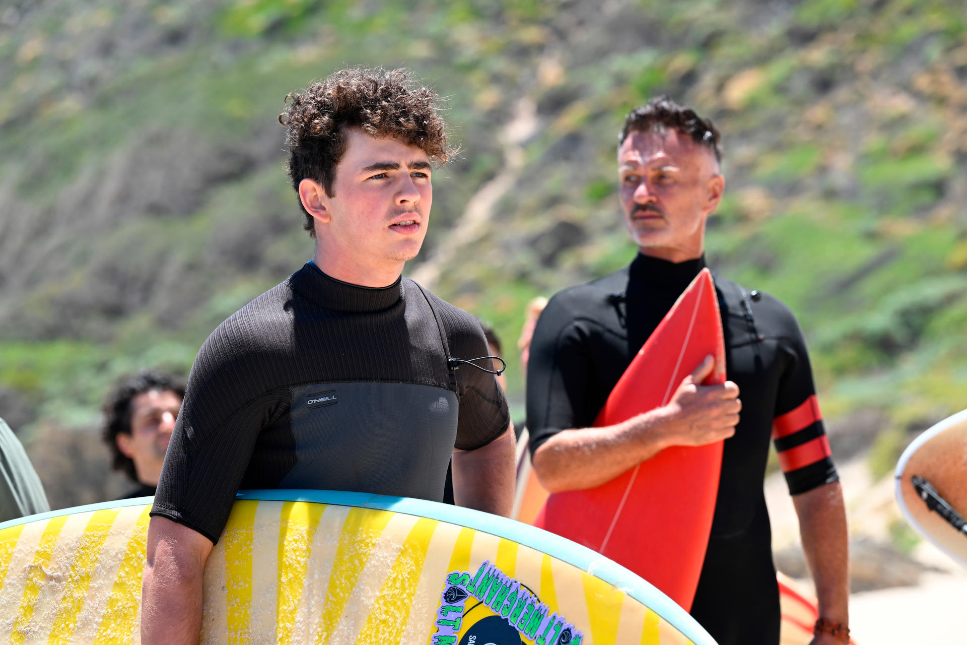 Junger Mann mit Surfbrett, ein anderer Mann im schwarzen Neoprenanzug hält rotes Brett. Hintergrund Strand.