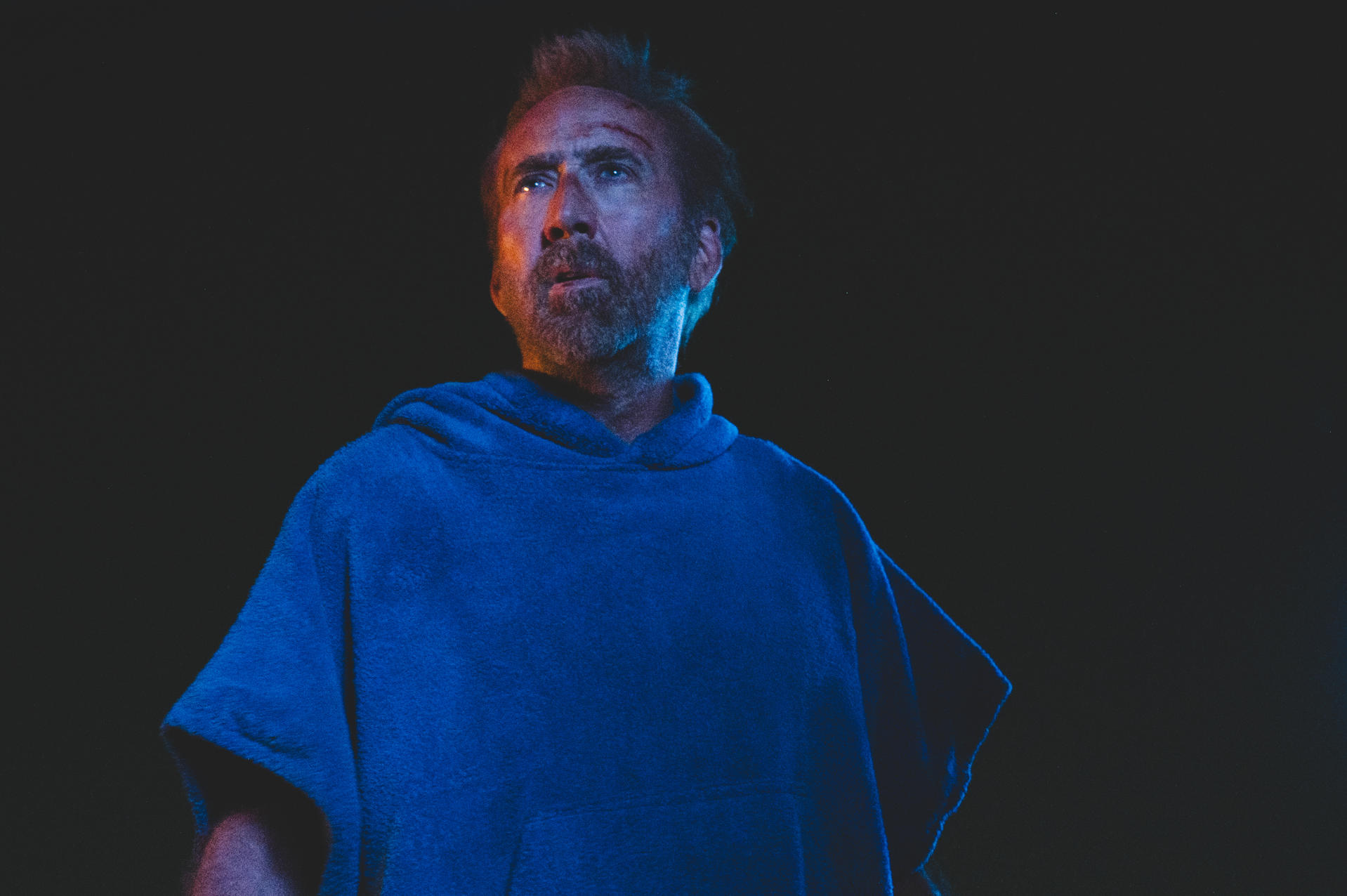 Nicolas Cage, nach oben blickend, trägt ein blaues, handtuchartiges Kleidungsstück, vor dunklem Hintergrund.