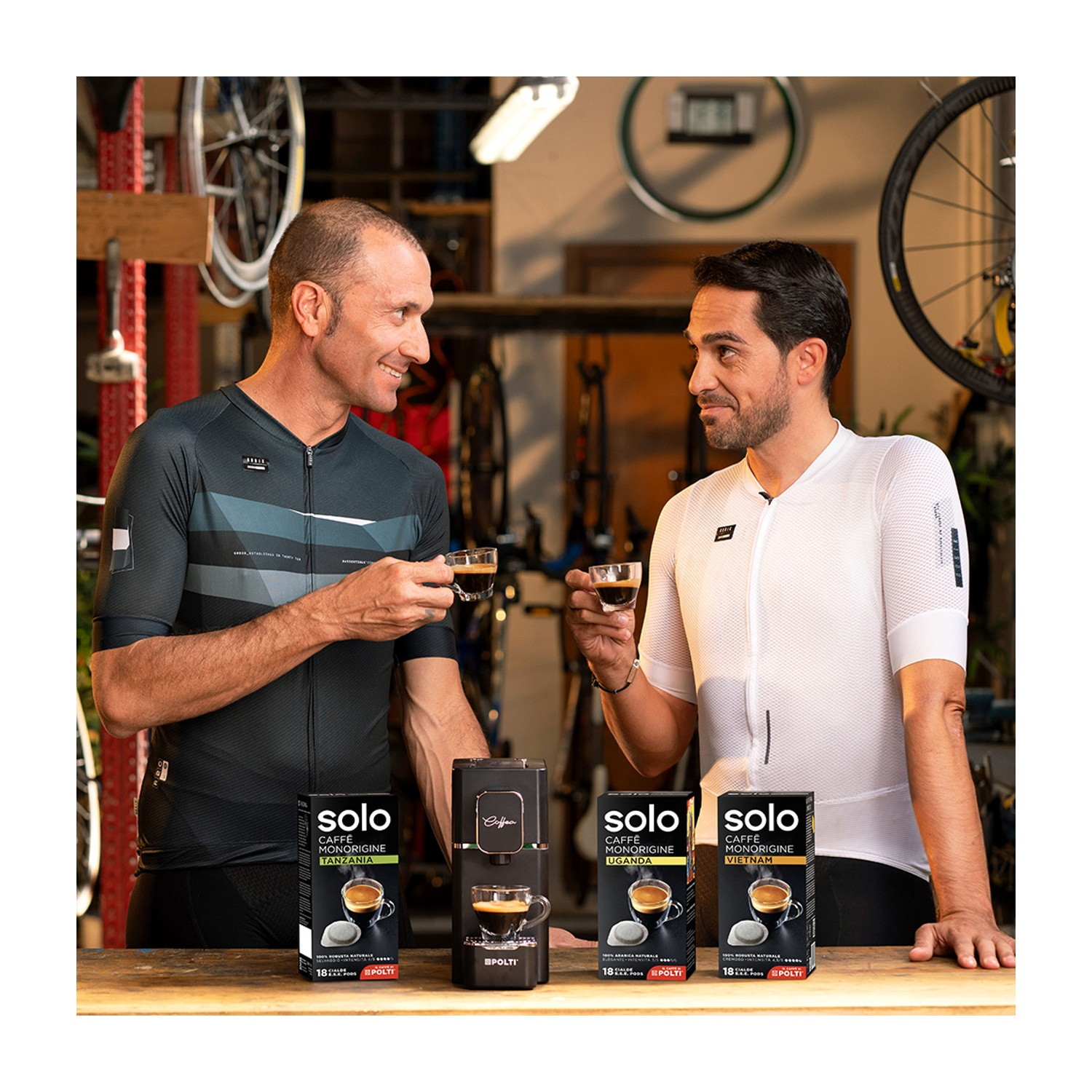 Due uomini con tazze di caffè, macchina per caffè Solo e cialde su un tavolo, negozio di biciclette sullo sfondo.