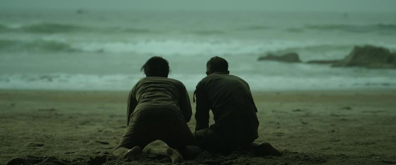Zwei Personen knien am Strand und blicken aufs Meer. Der Himmel ist bedeckt und das Wasser ist unruhig.