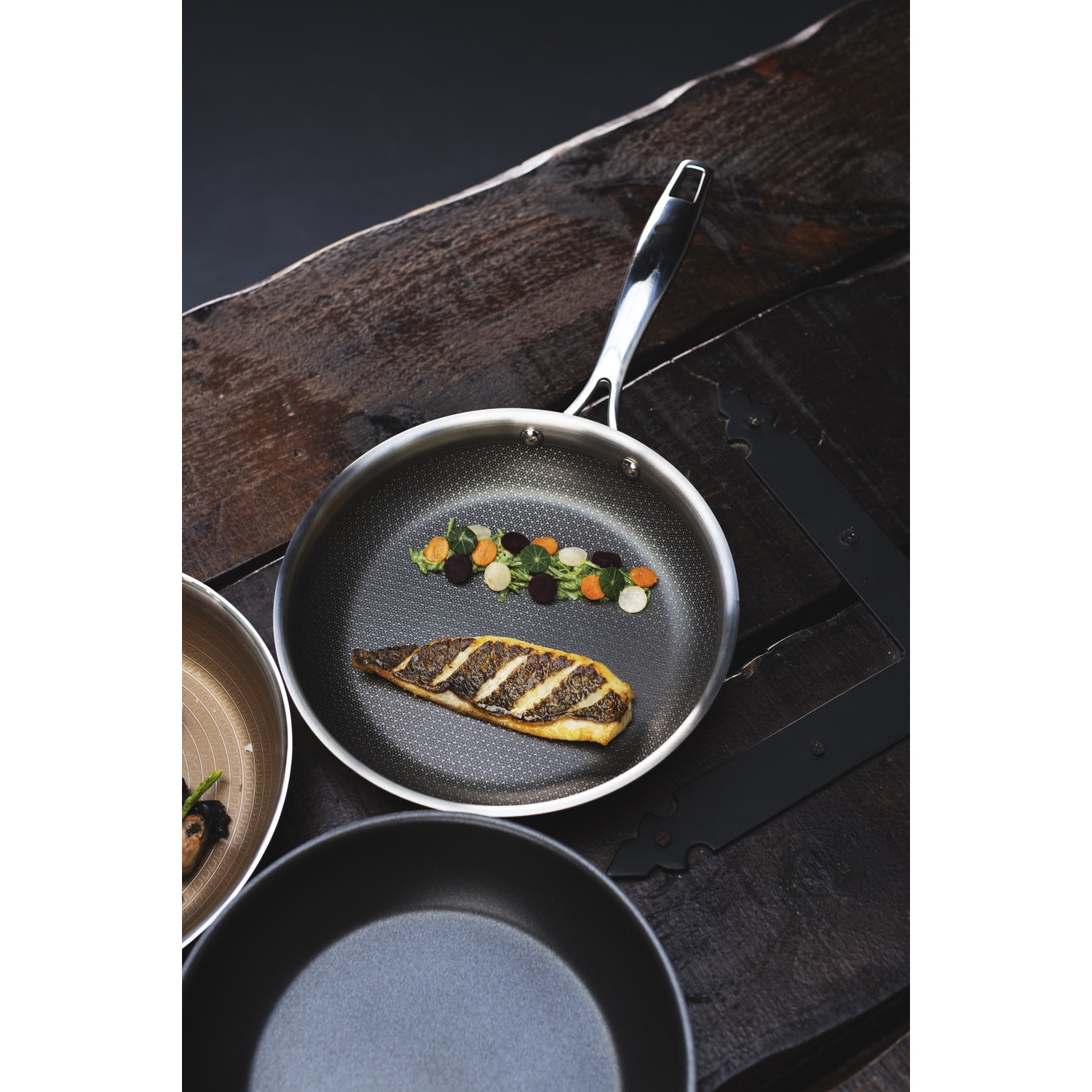 Una padella con pesce grigliato e verdure su un tavolo di legno. Altre padelle nelle vicinanze. Sfondo scuro.