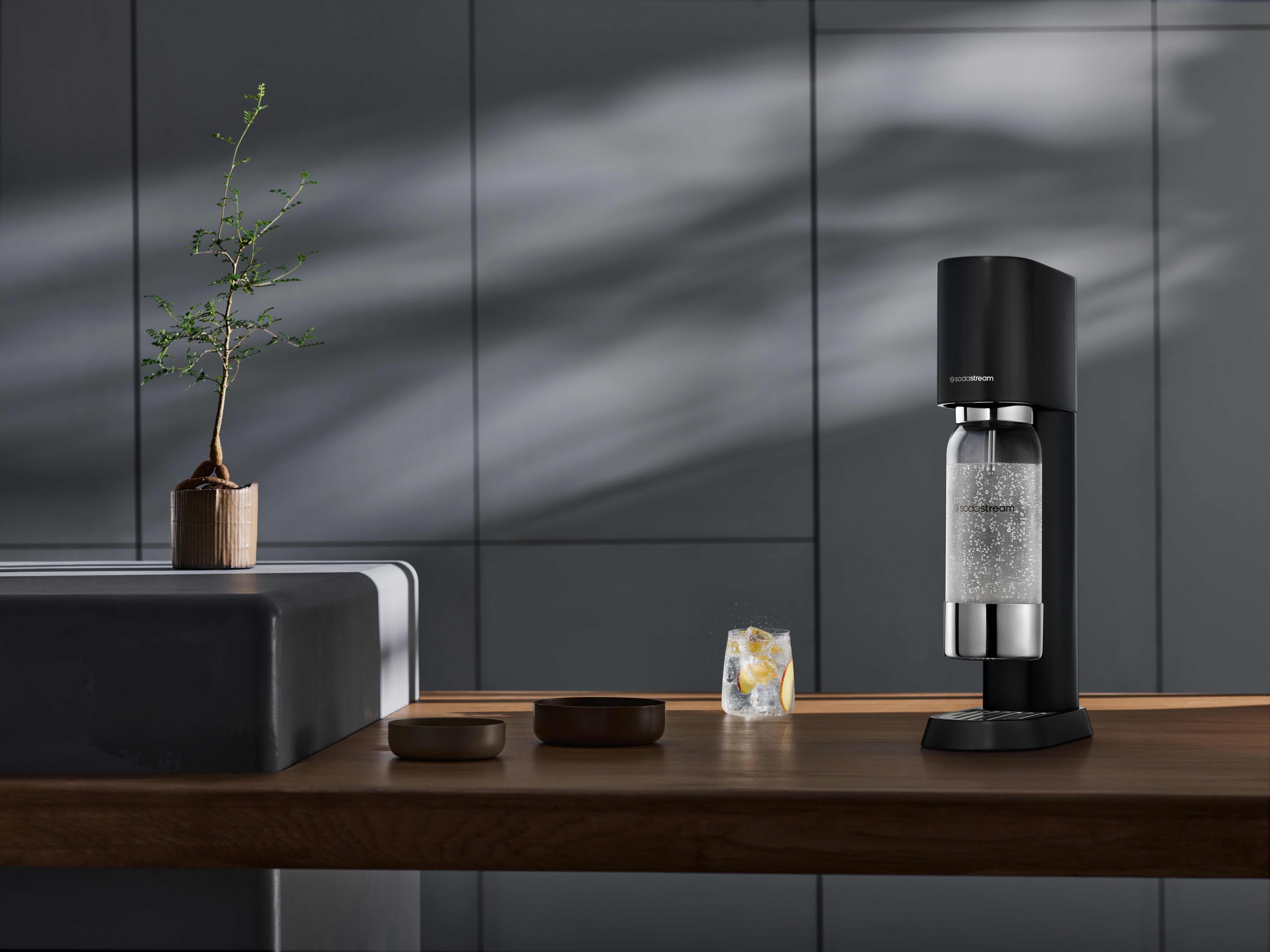 SodaStream-Maschine auf einem Holztisch mit einem Glas und Pflanzen. Die Maschine ist schwarz und silber.