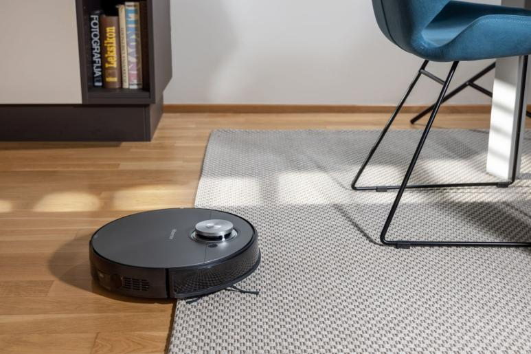 Egy robotporszívó fa padlót tisztít. Közel van egy dohányzóasztalhoz és egy kanapéhoz.
