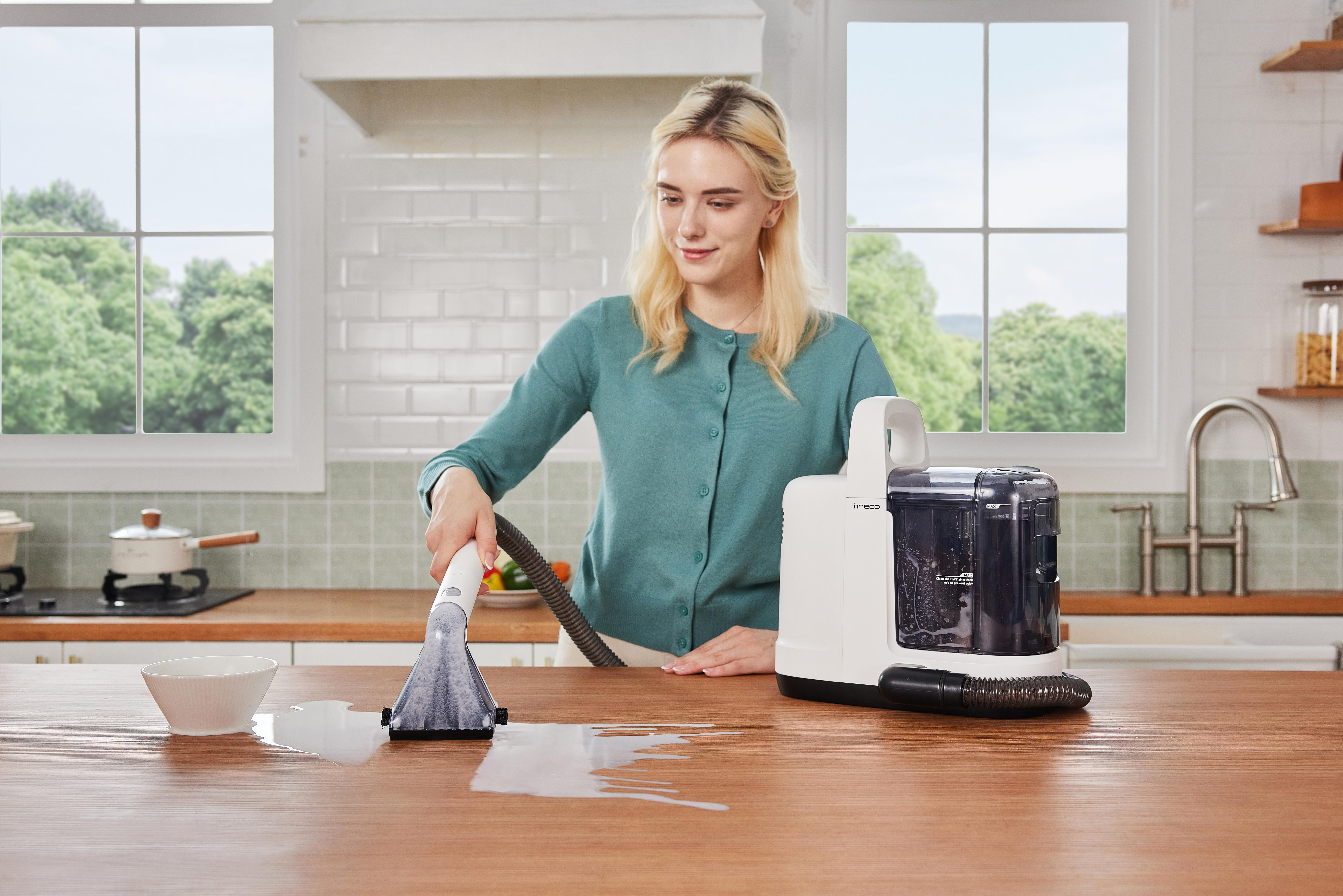 Eine Frau reinigt einen Holztisch mit einer Reinigungsmaschine. Eine Schüssel und verschüttete Flüssigkeit sind auf dem Tisch.