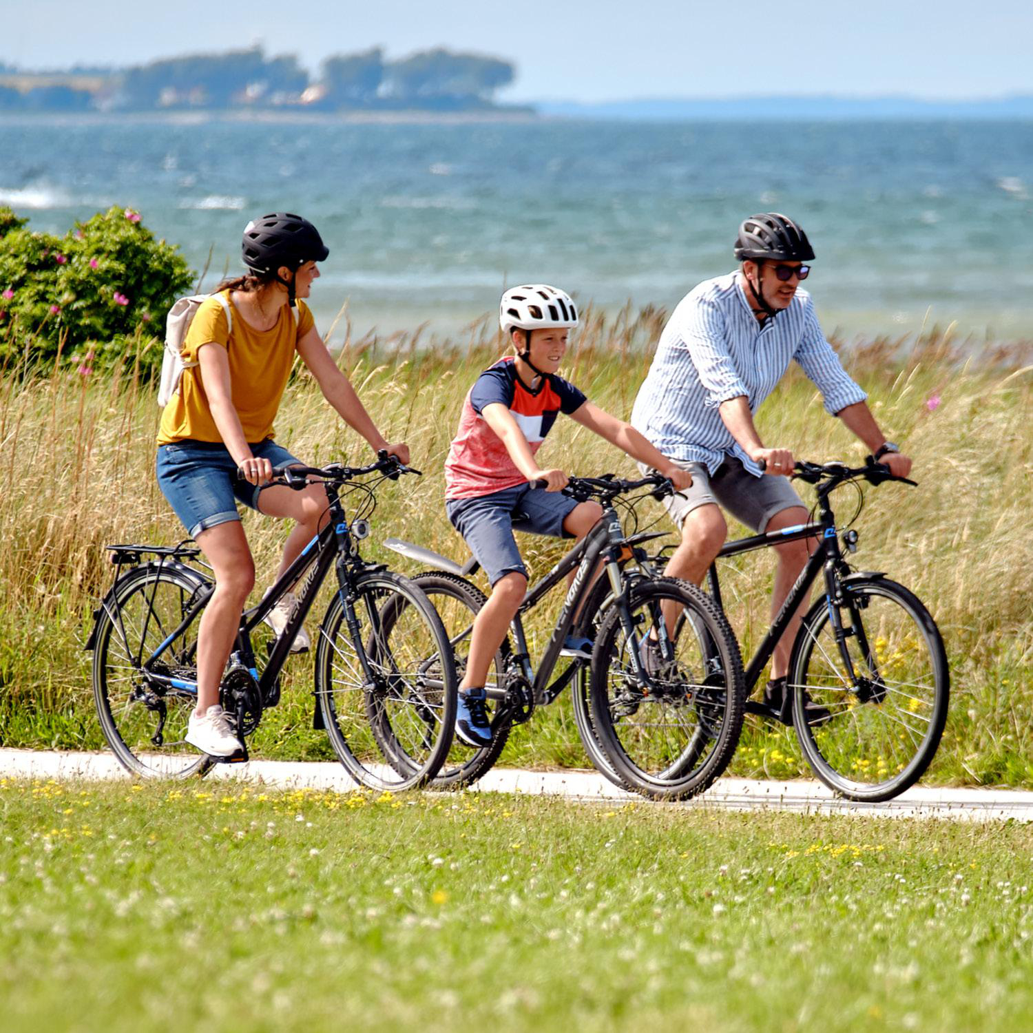 Dreiköpfige Familie fährt auf einem Weg Fahrrad. Das Meer ist im Hintergrund. Sonniger Tag.