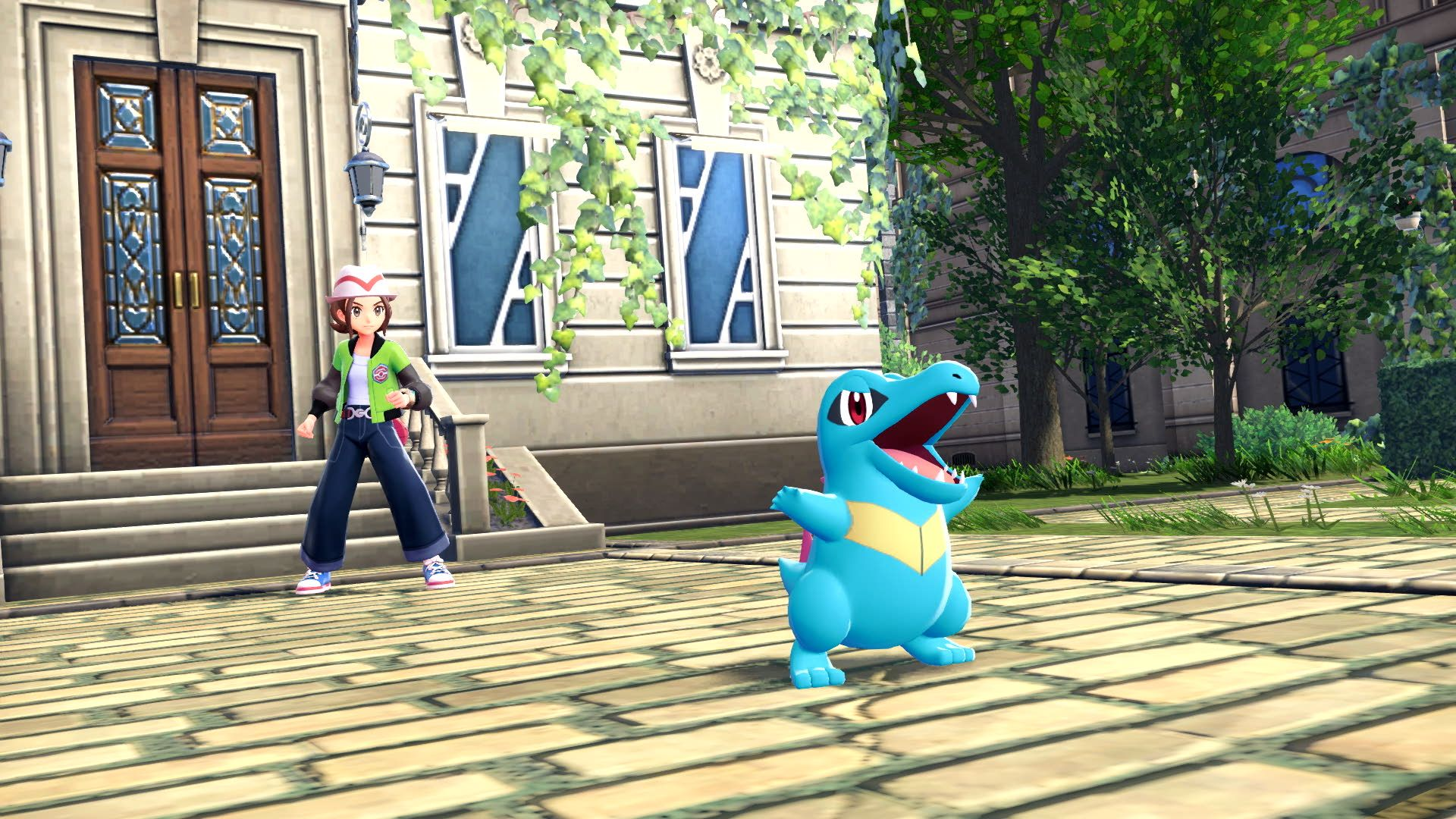 Un entraîneur se tient près d'un Pokémon bleu ressemblant à un crocodile. Bâtiment avec fenêtres et arbre en arrière-plan.