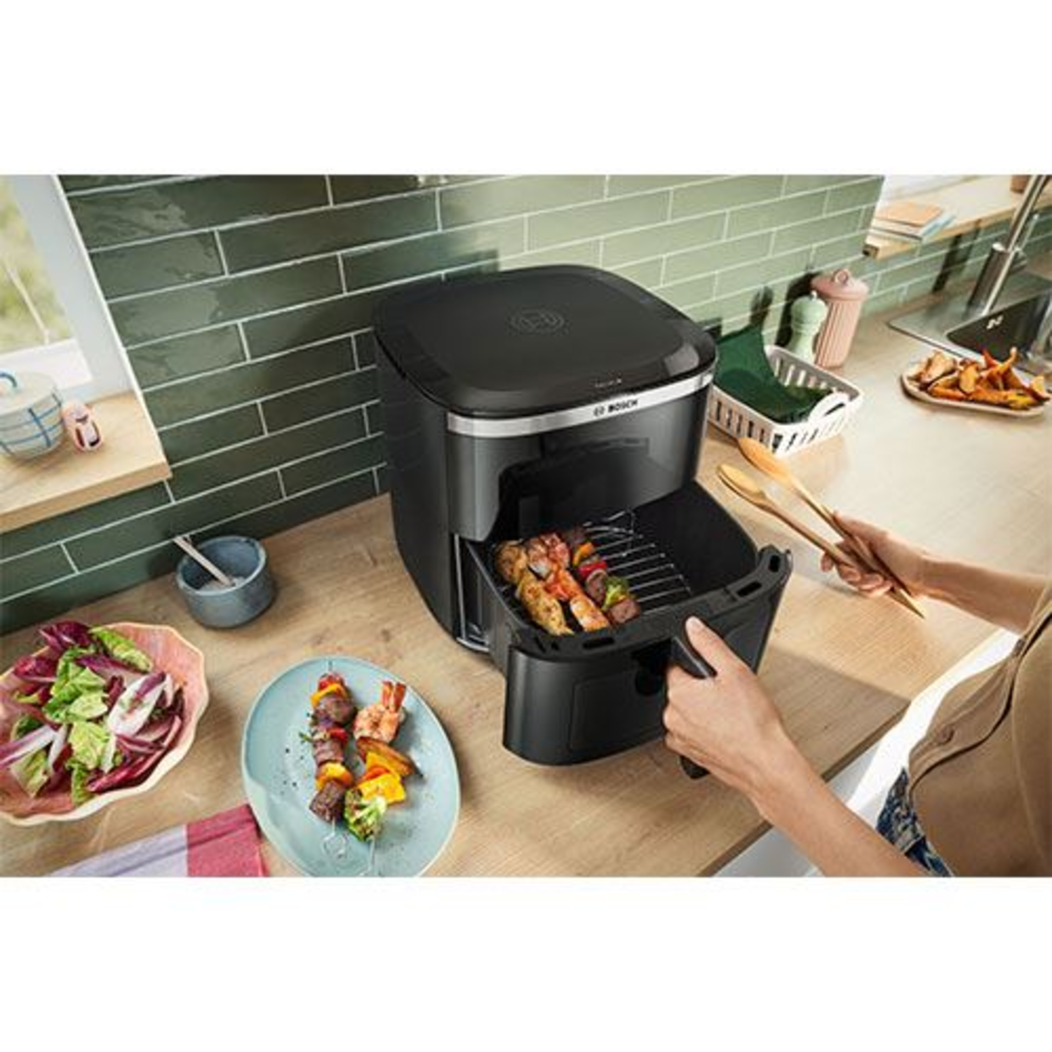 Une friteuse à air Bosch, noire, posée sur un comptoir. Une personne sort le panier avec de la nourriture.