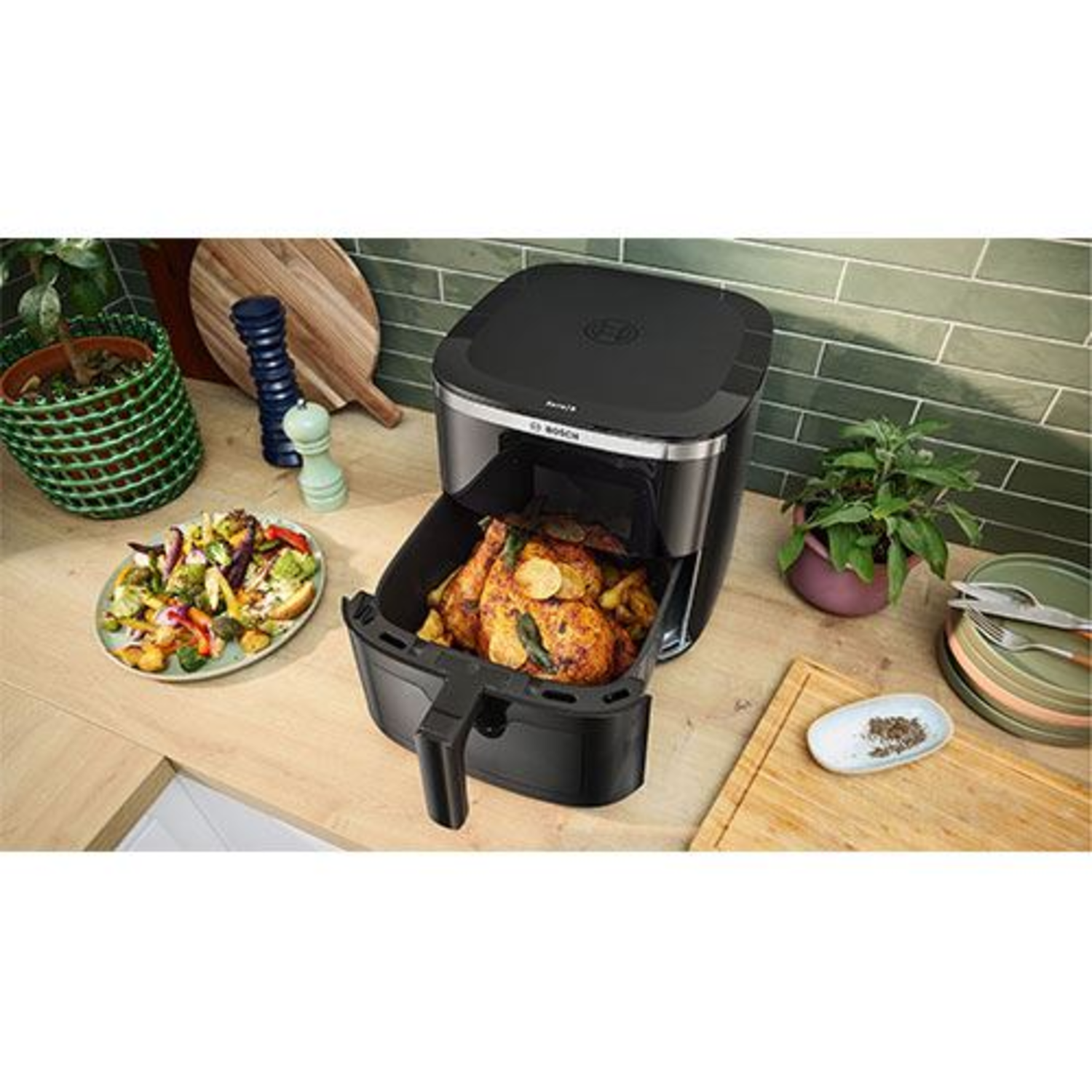 Friteuse à air avec poulet cuit, légumes et plantes sur un comptoir en bois.