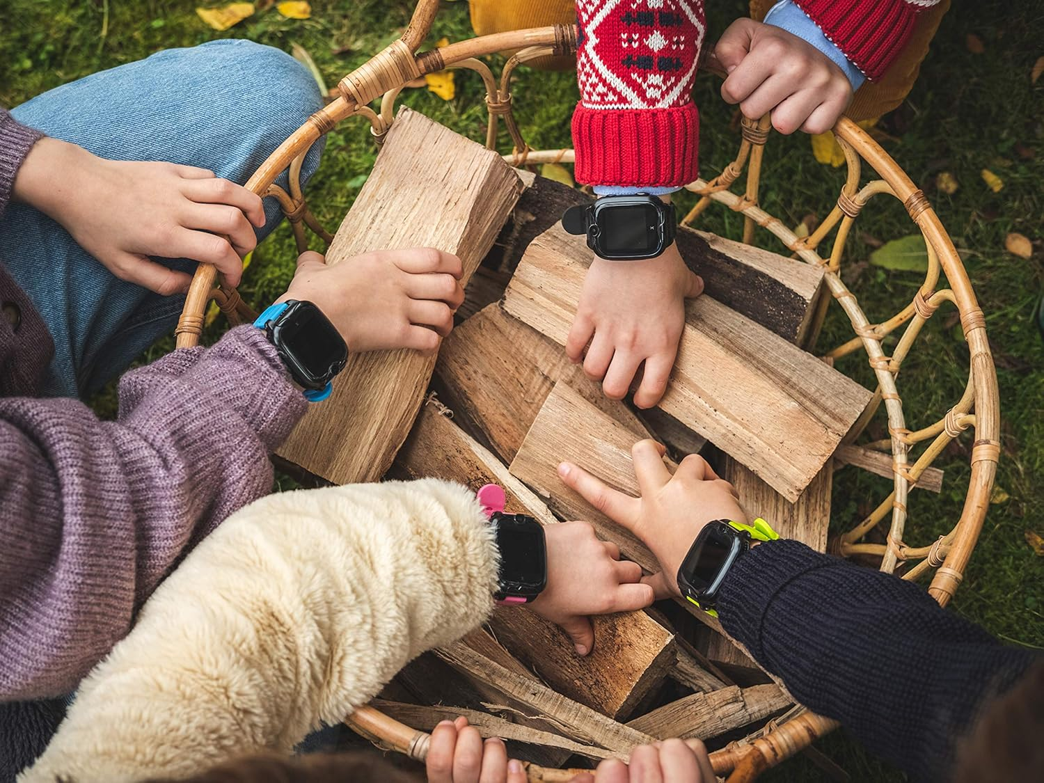 Manos de niños alrededor de una cesta llena de leña, usando smartwatches con correas de diferentes colores.