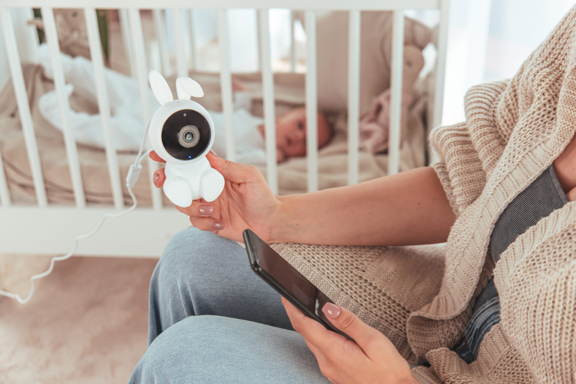 Kobieta trzymająca monitor dziecięcy w kształcie królika i telefon. Dziecko jest w łóżeczku w tle.