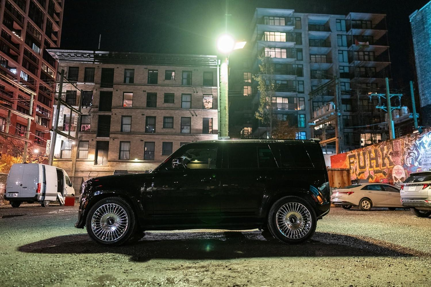 Ein schwarzer SUV parkt nachts. Gebäude und eine Graffiti-Wand sind im Hintergrund.