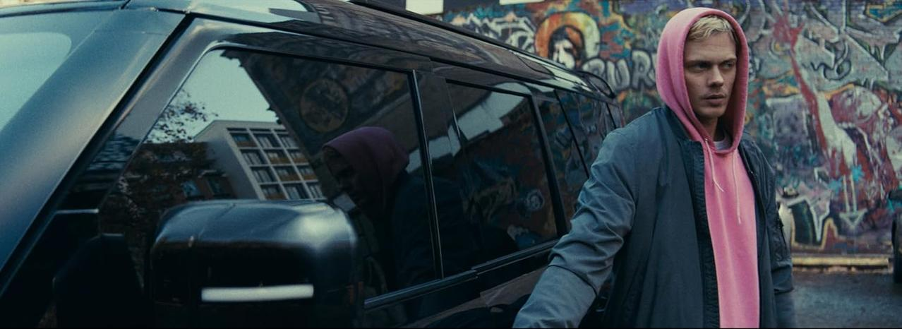 Ein Mann in einem pinken Kapuzenpullover lehnt an einem schwarzen Auto, mit einem bunten Graffiti-Hintergrund.