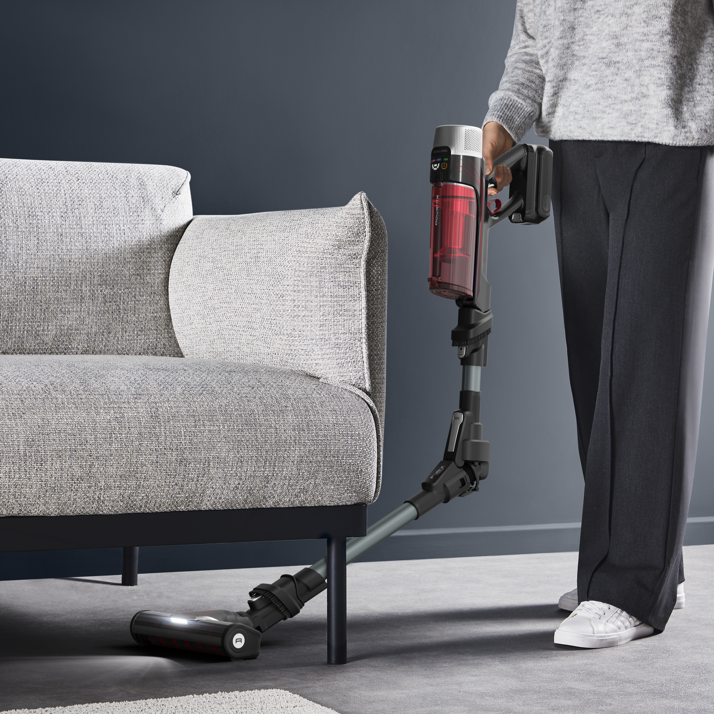 Kobieta odkurza jasnoszarą sofę czerwono-czarnym bezprzewodowym odkurzaczem. Ciemnoniebieska ściana.